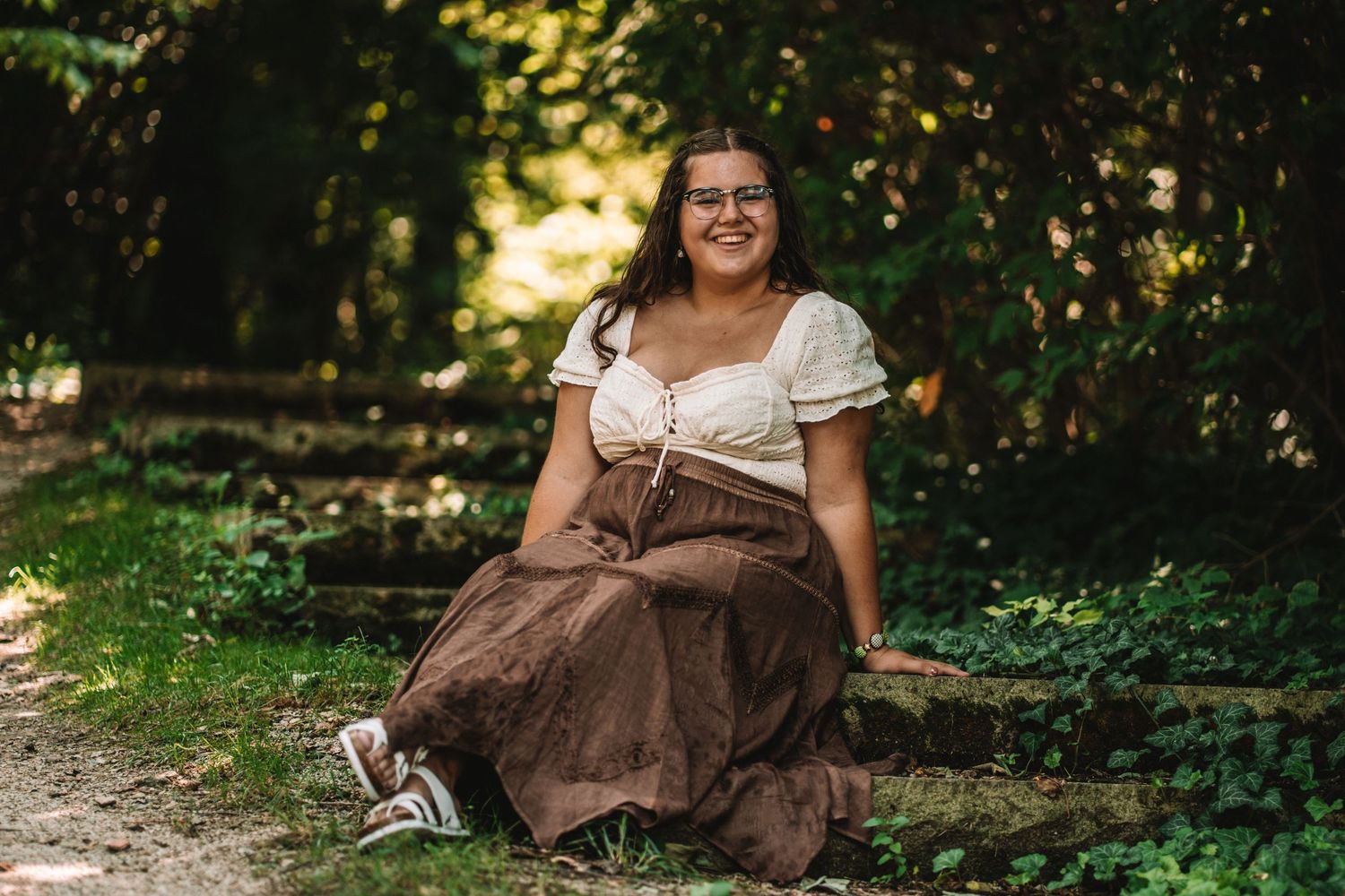 Person in brown dress and cream top posing outdoors against natural foliage background.