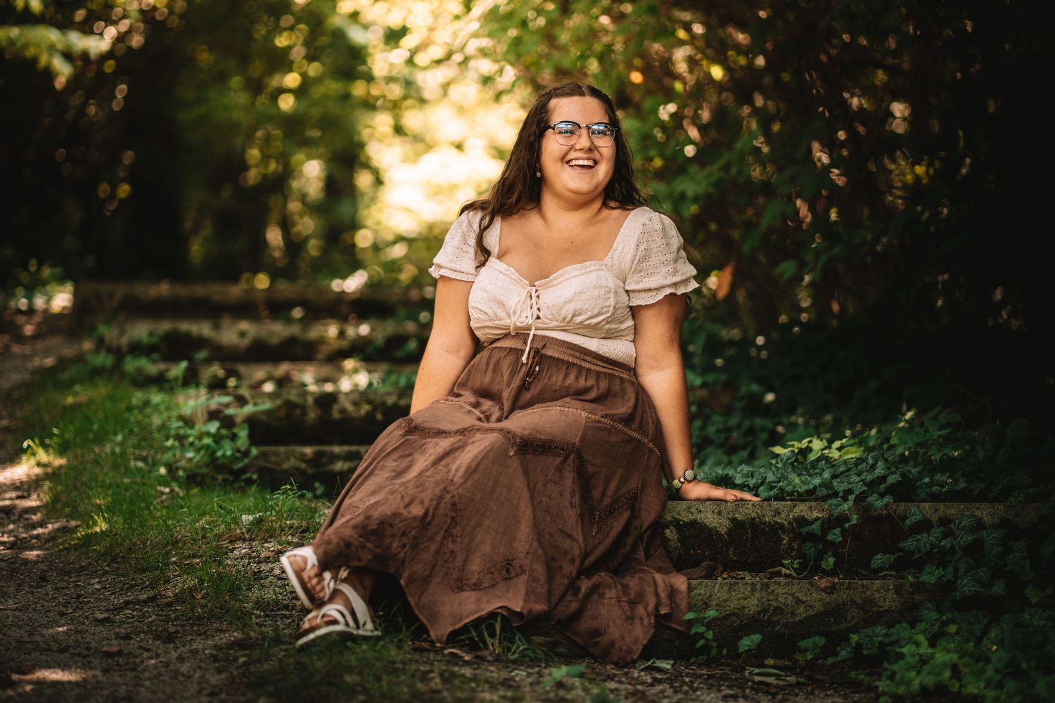 Reading peacefully in a brown dress against a natural forest backdrop with dappled sunlight.