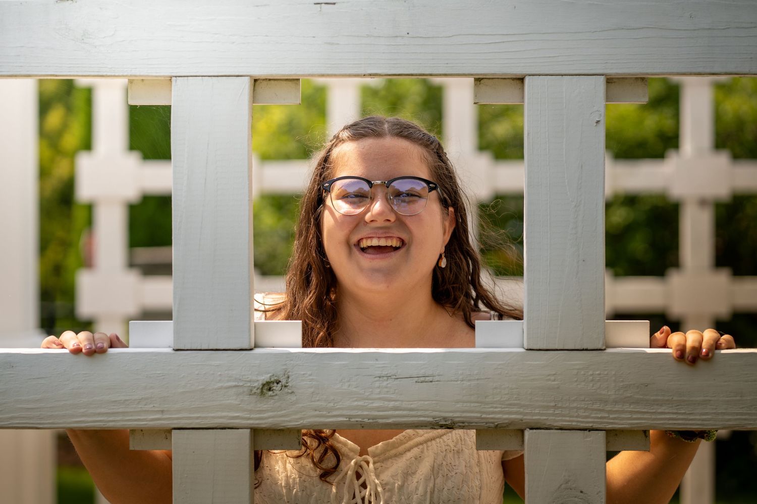 A joyful and candid expression peeks through white fence slats in this bright outdoor photography moment.