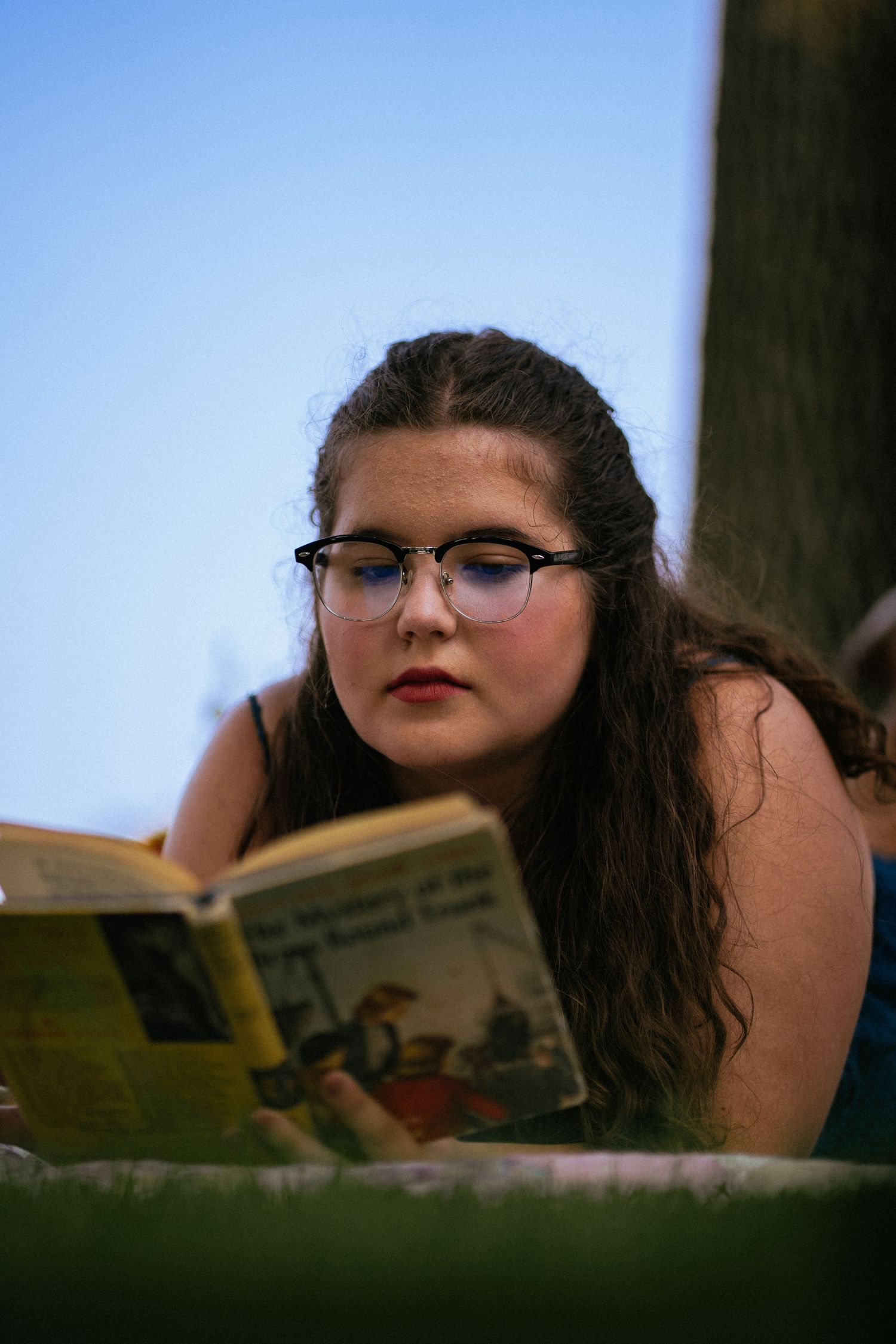 A close-up portrait of someone absorbed in reading a book against a bright blue sky background.
