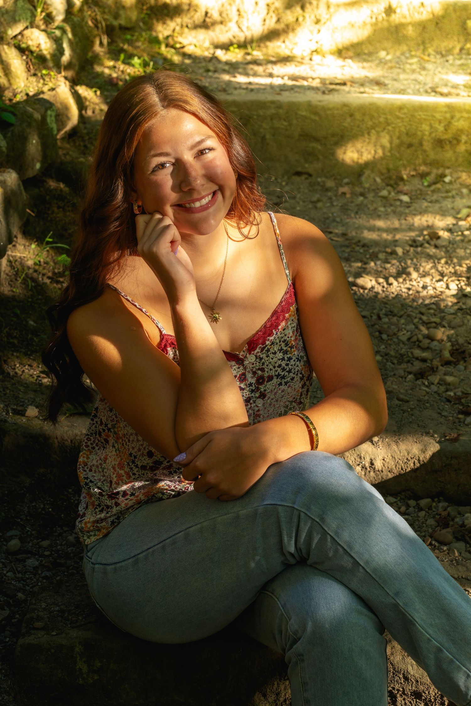 Person sitting outdoors in dappled sunlight wearing jeans and a dark patterned top.