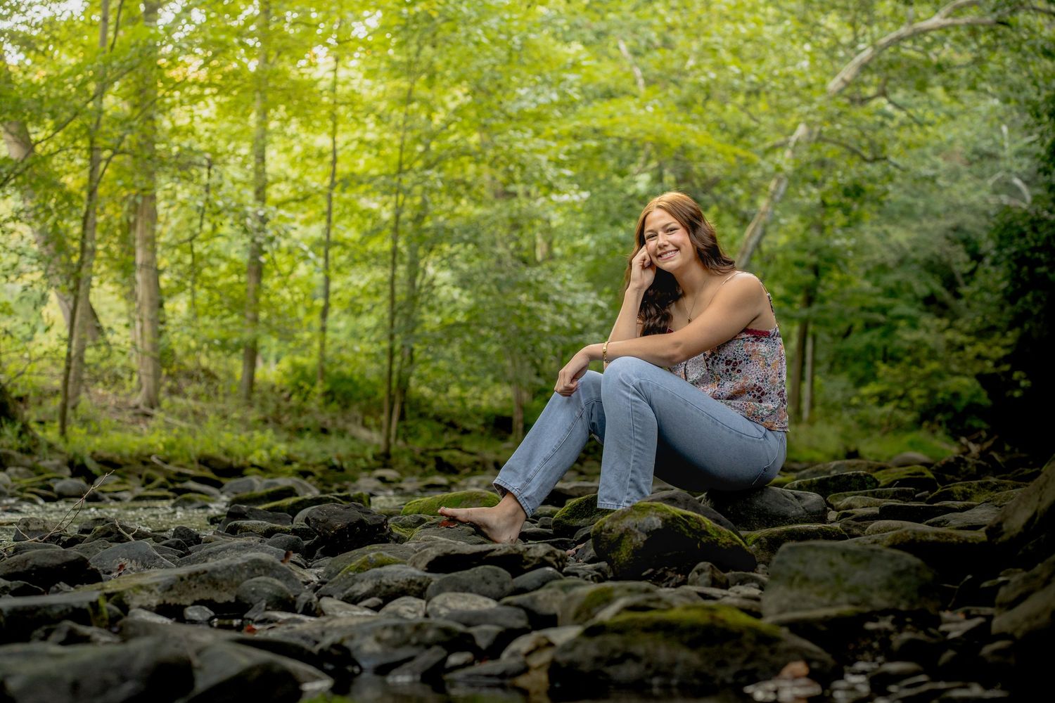 A person in blue jeans sits thoughtfully on a stone wall surrounded by lush green forest scenery.