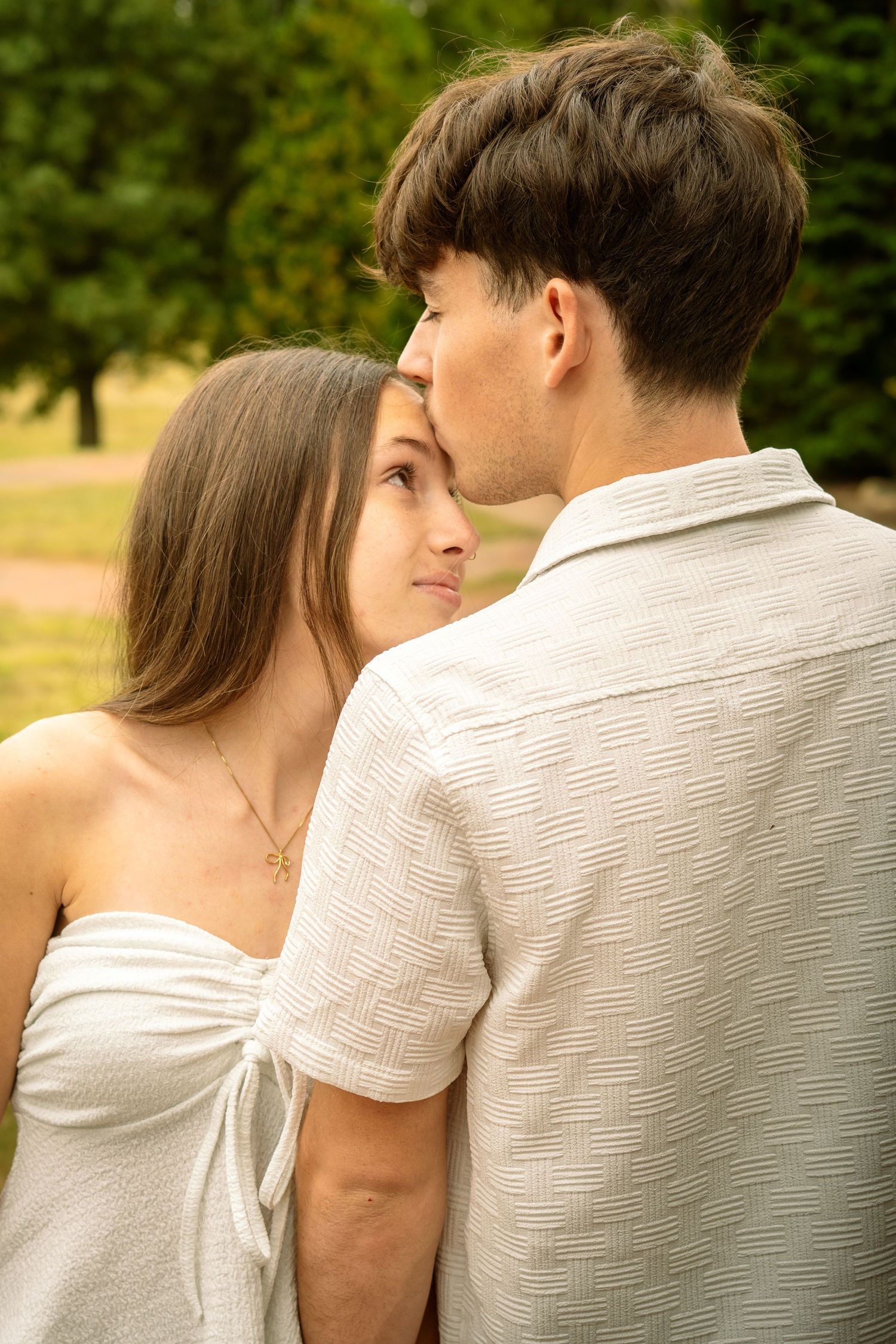 A romantic couple in white outfits share an intimate moment outdoors in a sunny park setting.