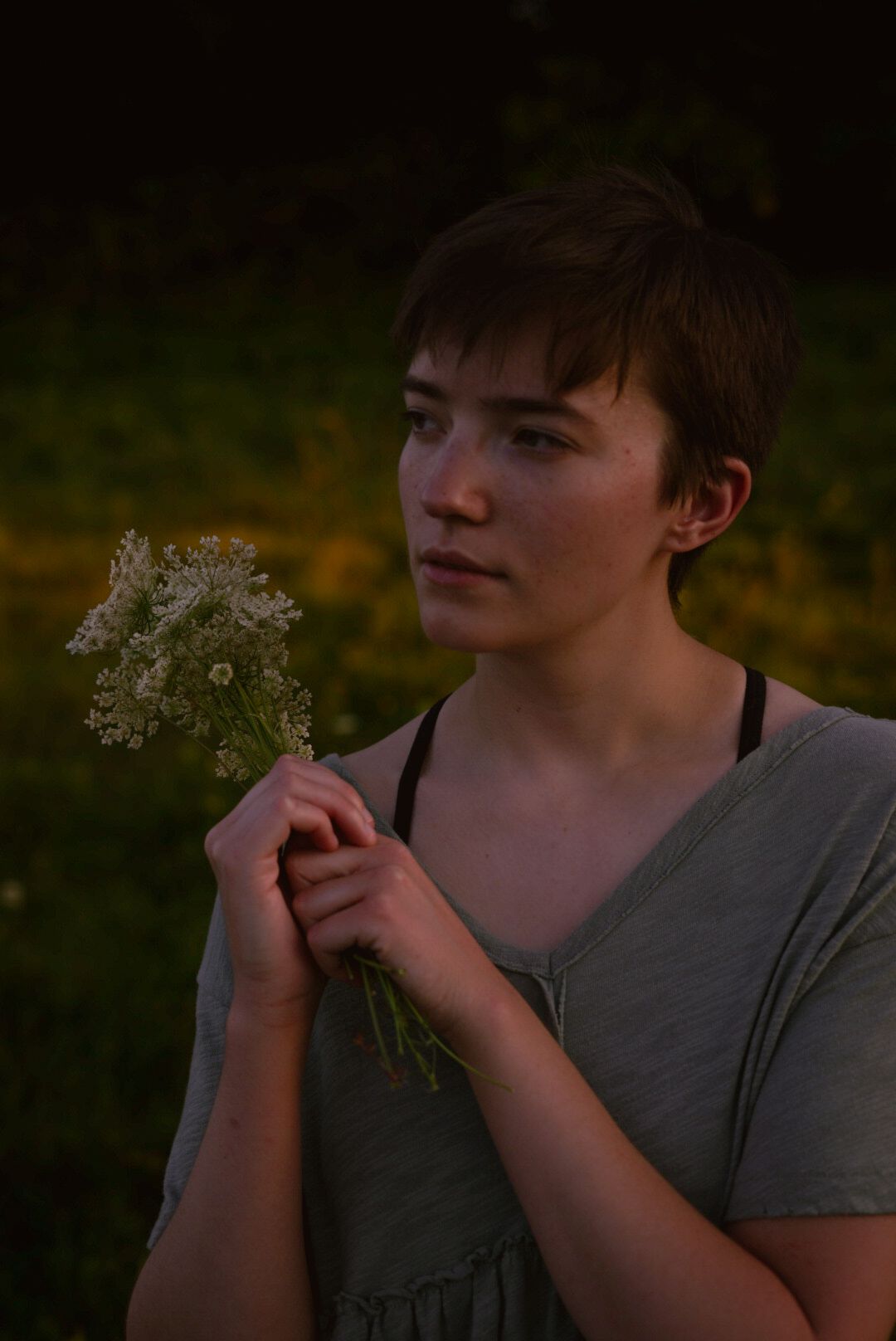 a girl staring out into the trees while holding a bouquet of wildflowers. trees are behind her as well with yellow flowers in the field