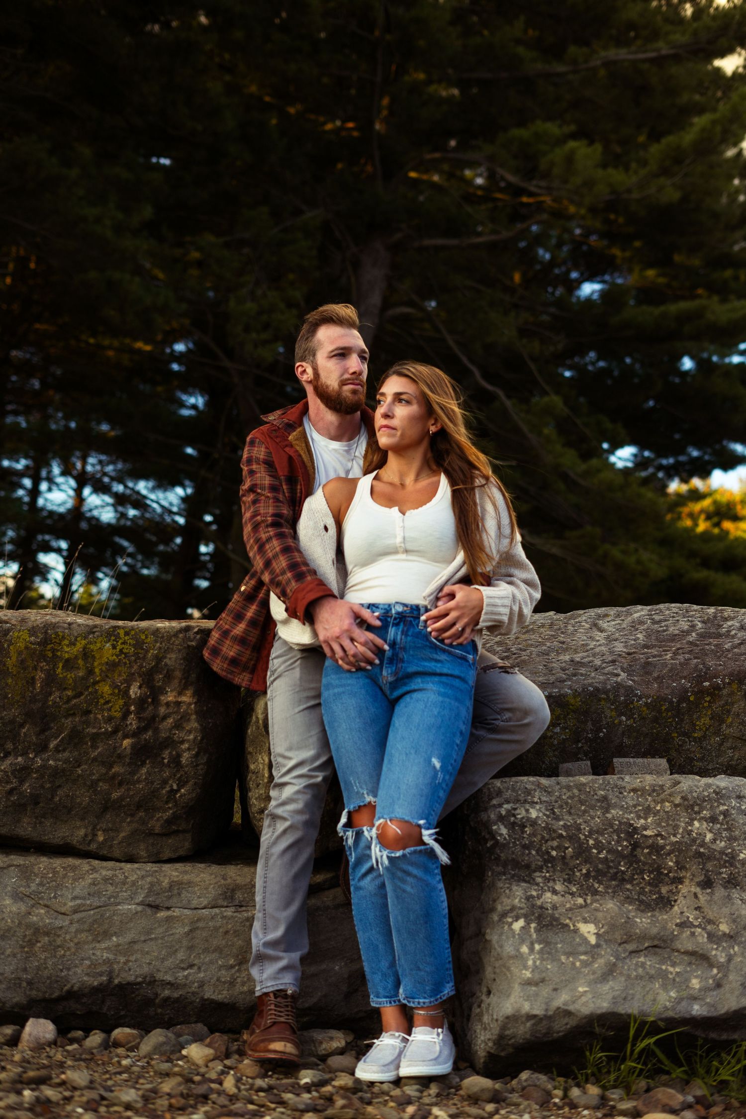 a man and woman standing on a Rocky shore. looking in opposite directions. pine trees loom behind them