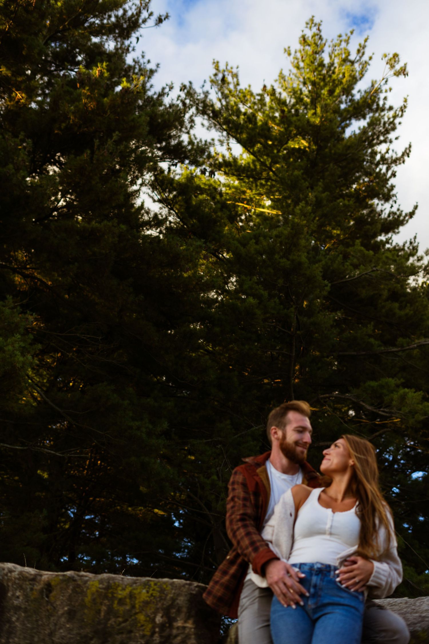 a man wrapping arms around fiancees waist. they are smiling and looking at each other. blue skies and green pine trees standing tall behind