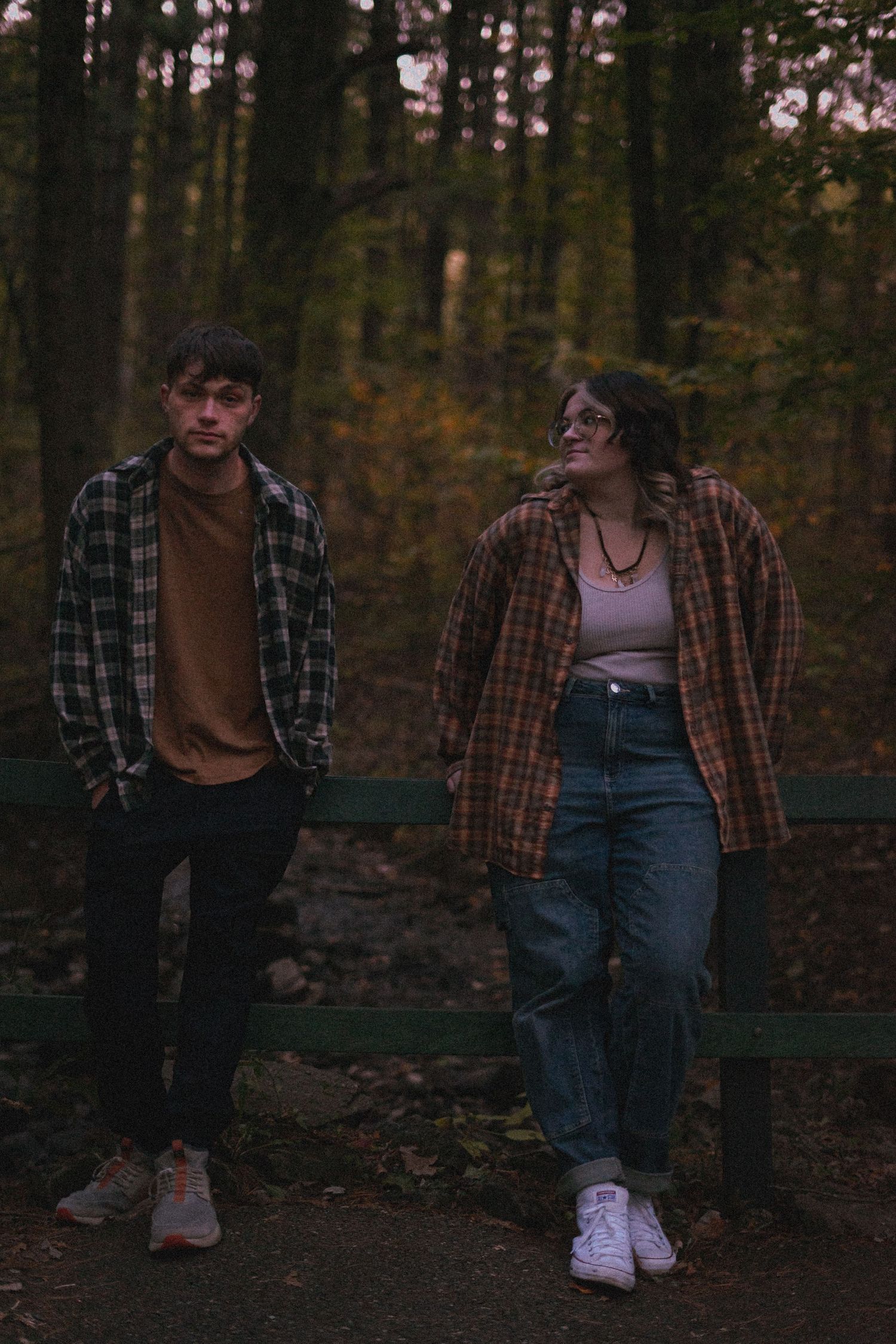 a woman smiling and looking at her boyfriend while he smiles into the camera. both wearing flannels and on a bridge.