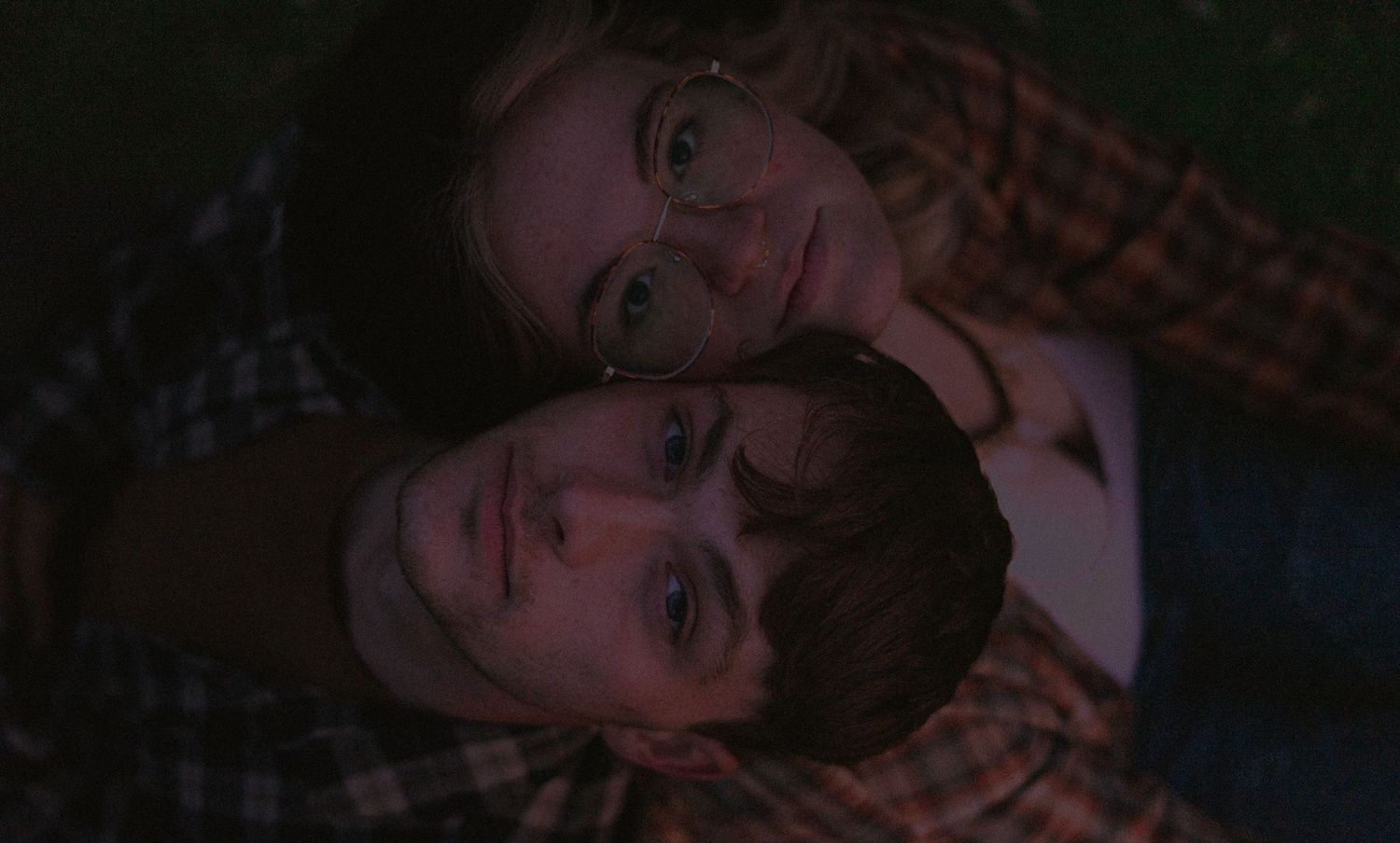 a couple leaning against each other, ones head on the others shoulder. starting up at the camera while sitting on the ground in the field