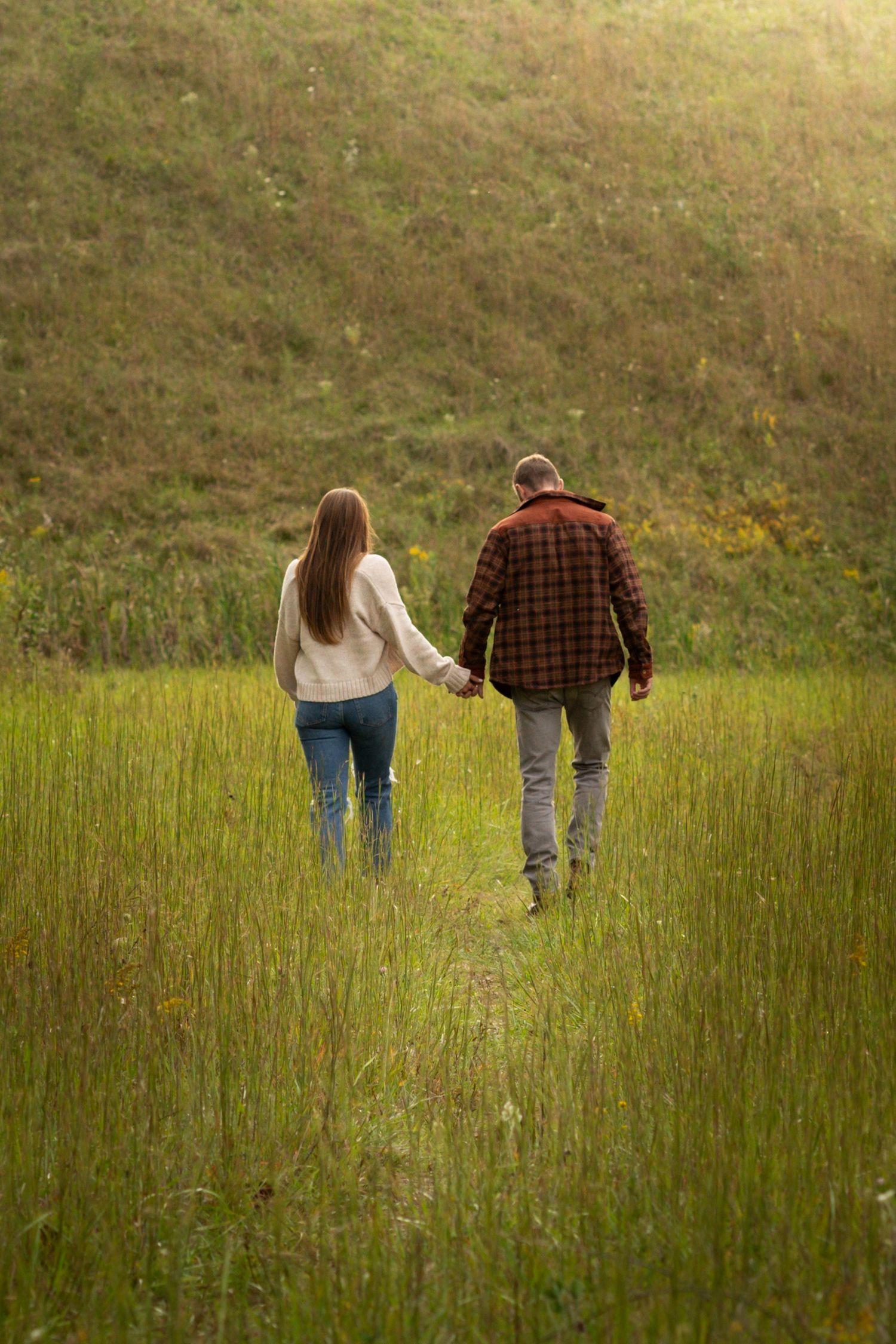 happy couple holding hands and walking through a green field of grass. a big hill is ahead of them and the sun is shining from above