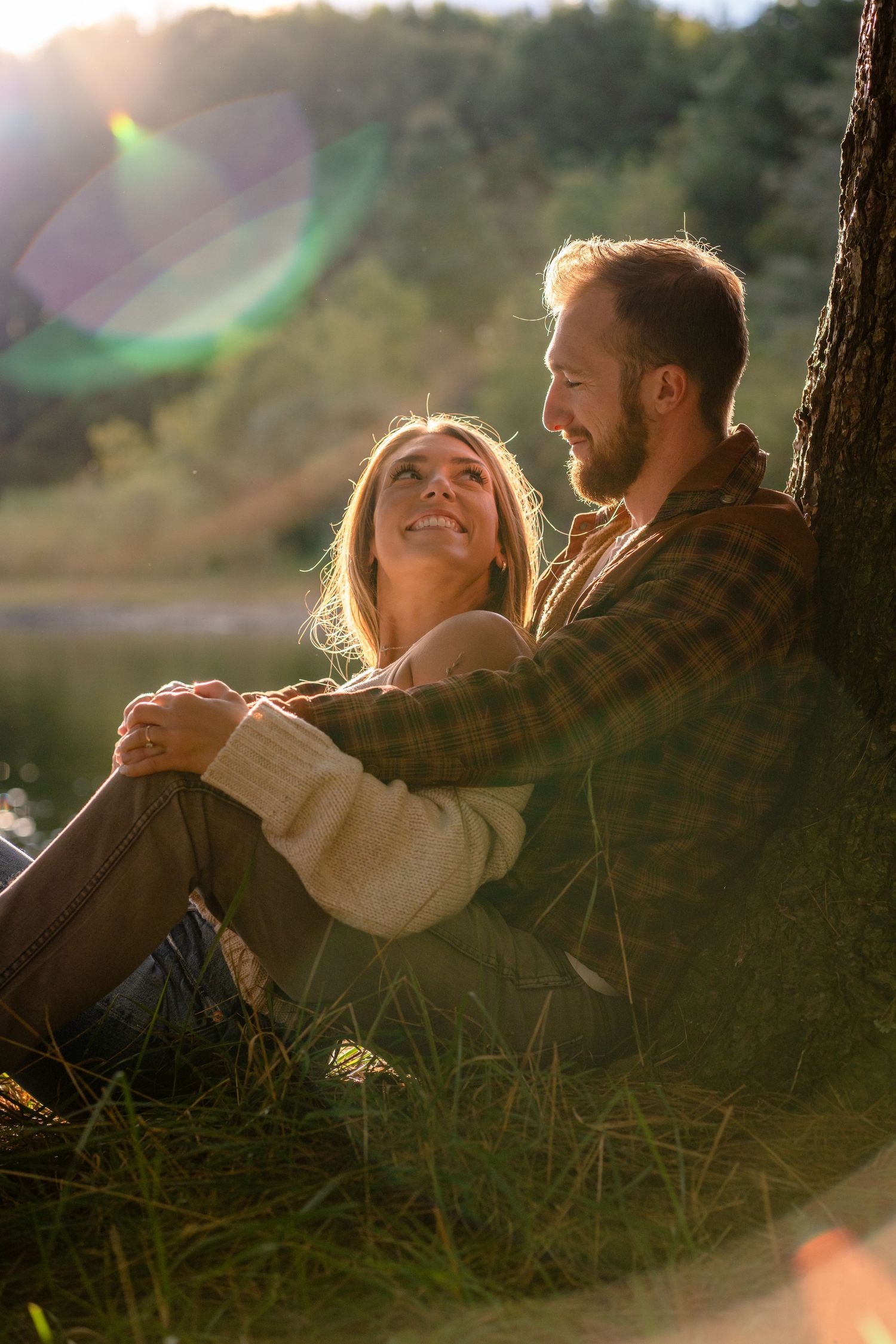 a happy couple laughing and smiling at each other while taking engagement photos. both are leaning against a tree and sitting on the ground