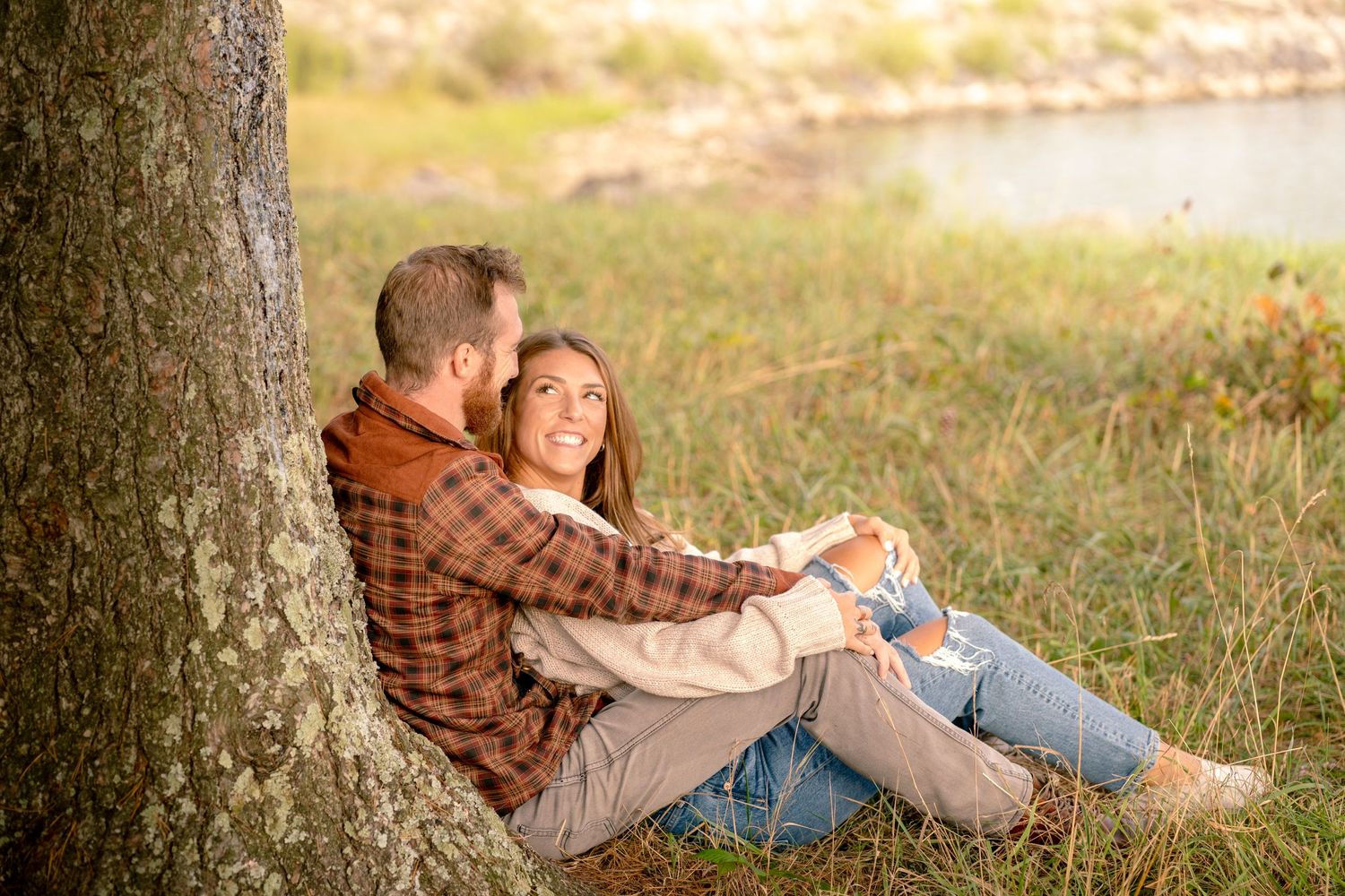 a happy couple laughing and smiling at each other while taking engagement photos. both are leaning against a tree and sitting on the ground