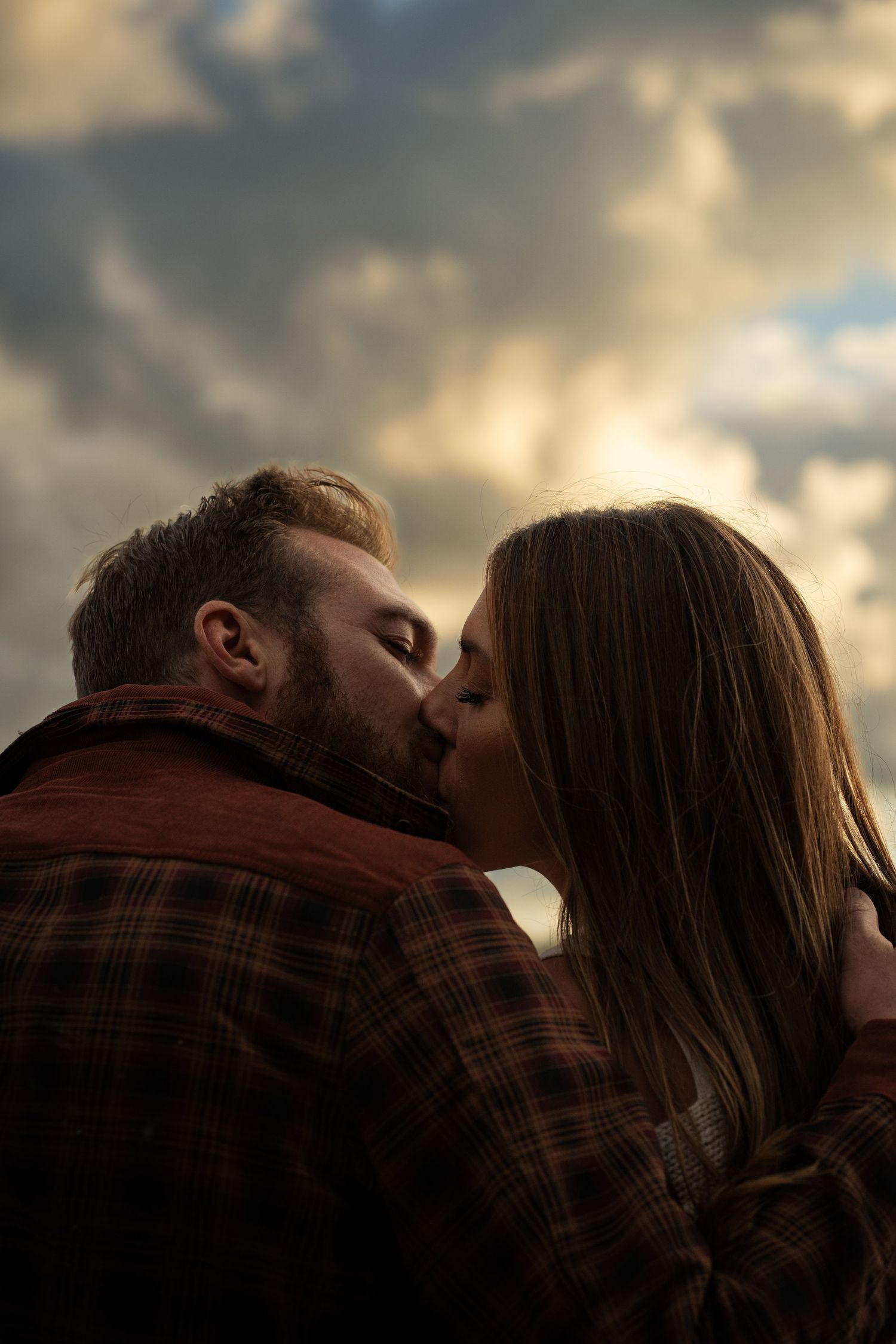 an engaged couple kissing with the sky glowing gold with golden clouds as the sun tries to Peirce through. Engagement Photgraphy