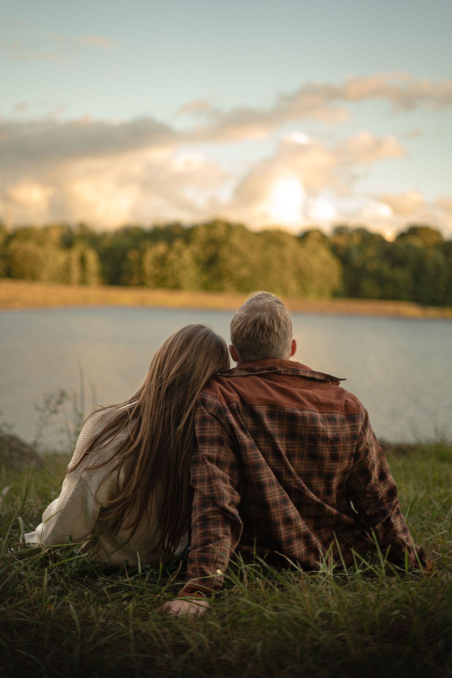 a man and woman sitting and holding hands in green grass at the edge of the water, watching the sun glow as it sets behind the pine trees