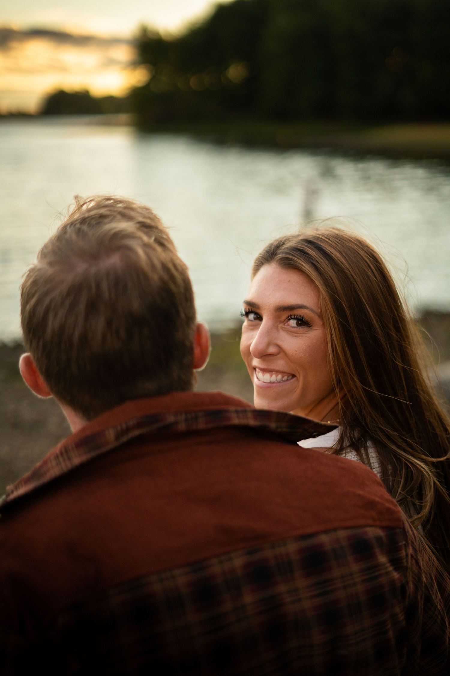 a fiancee looking over her fiances shoulder, smiling. sitting on Lake shore while the sun shines and bounces off water in the background.