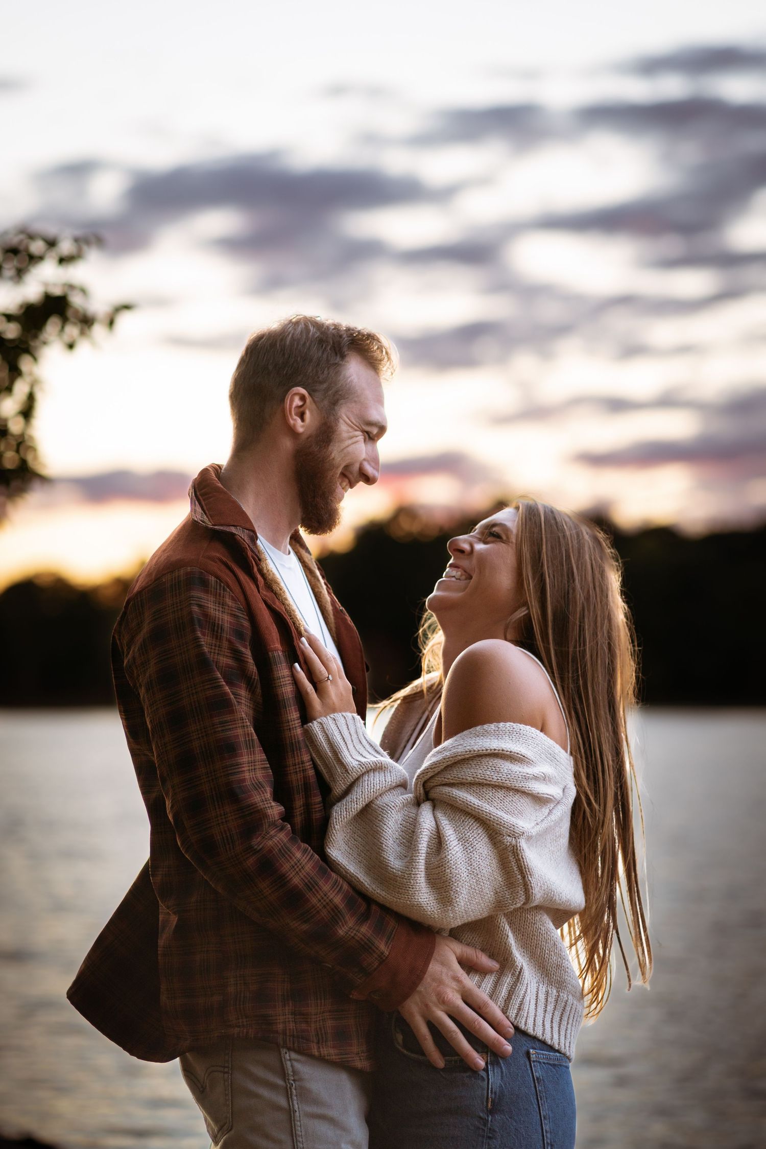 a man and woman standing lakeside, embracing each other. smiling and laughing while the sun begins to set over the trees behind them