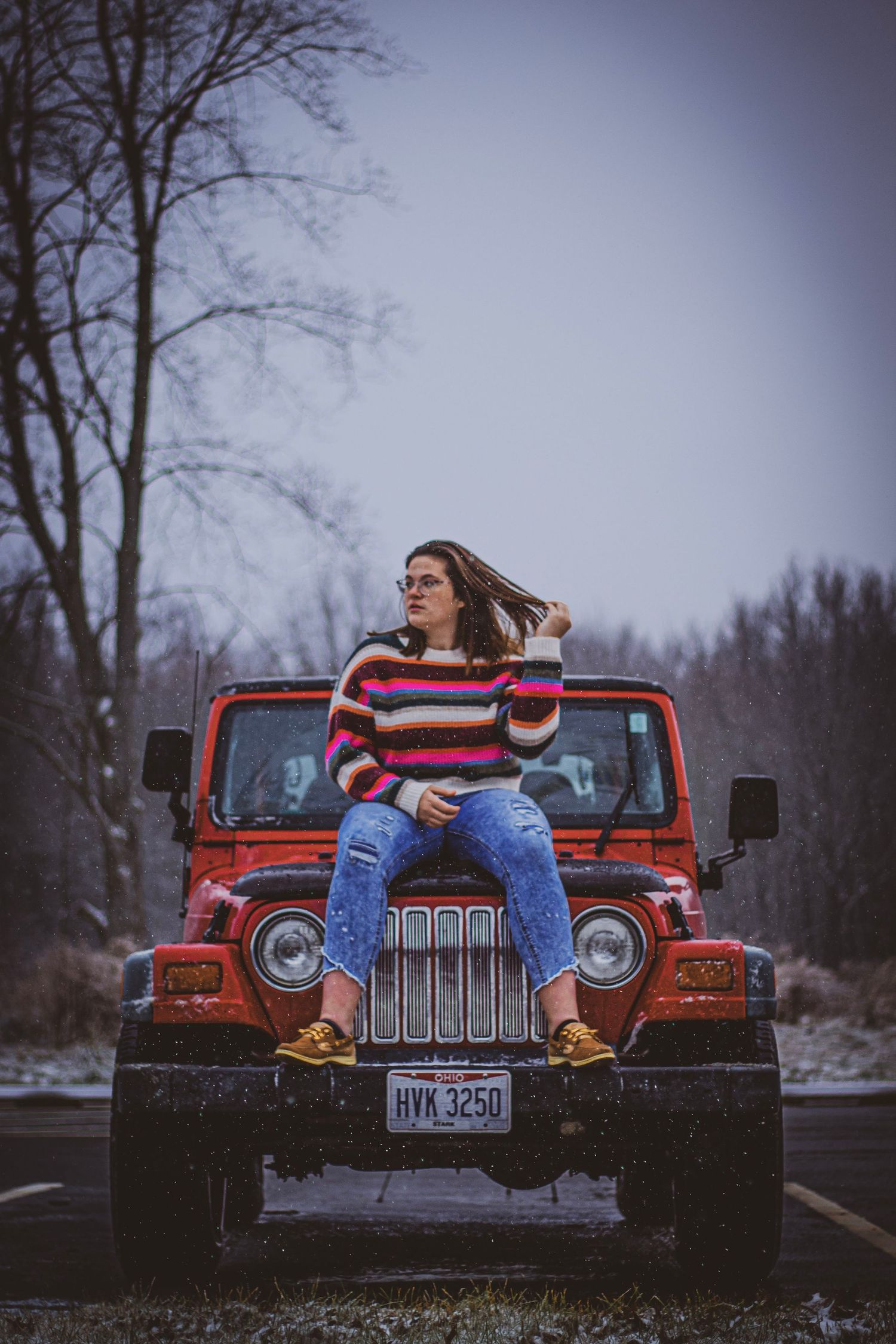 a woman in a bright, striped sweater, sitting on the hood of a red jeep while it snows. the white snow is falling from the sky