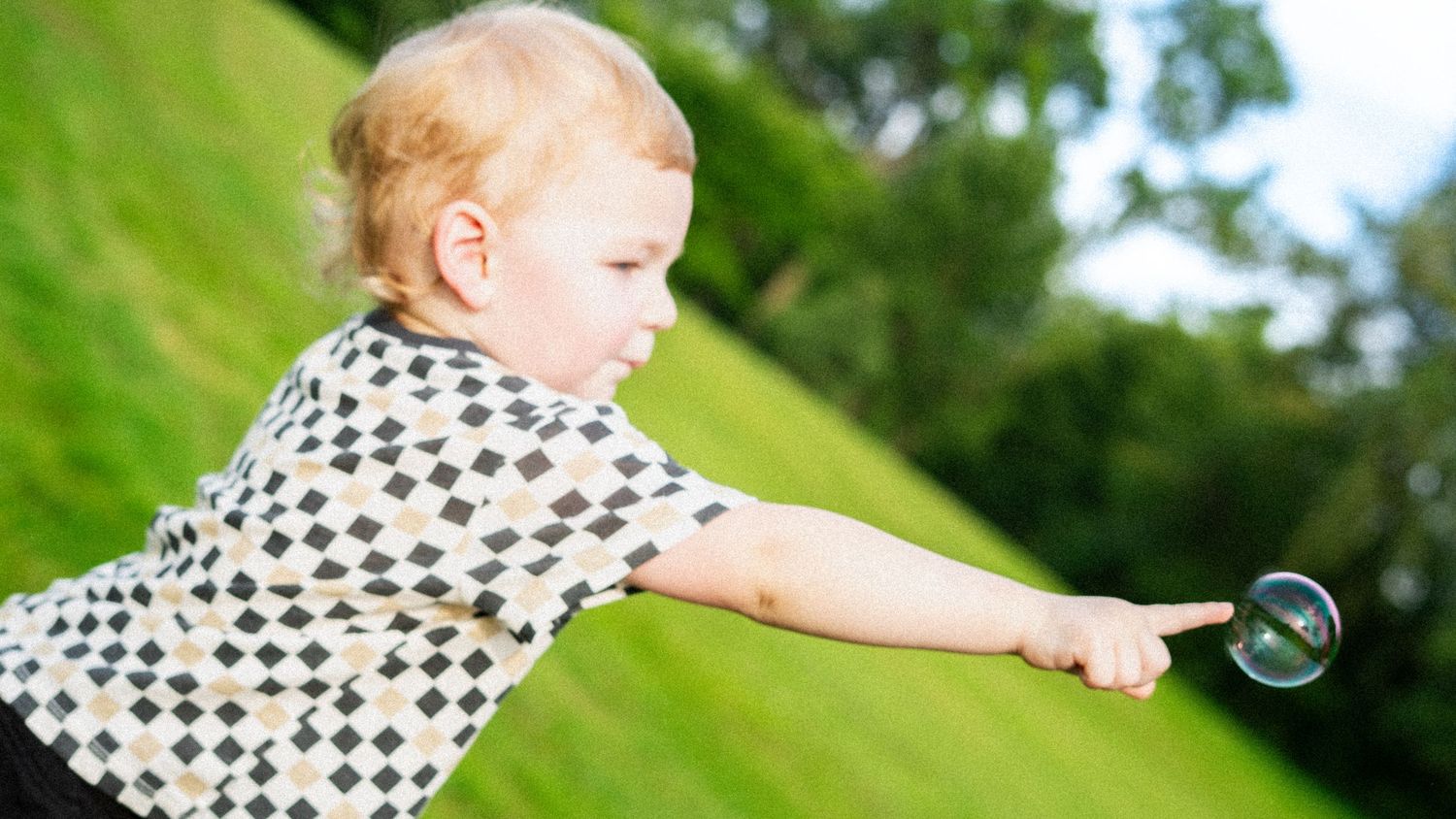 Toddler in checkered shirt reaches for floating soap bubble in sunny outdoor setting with green background.