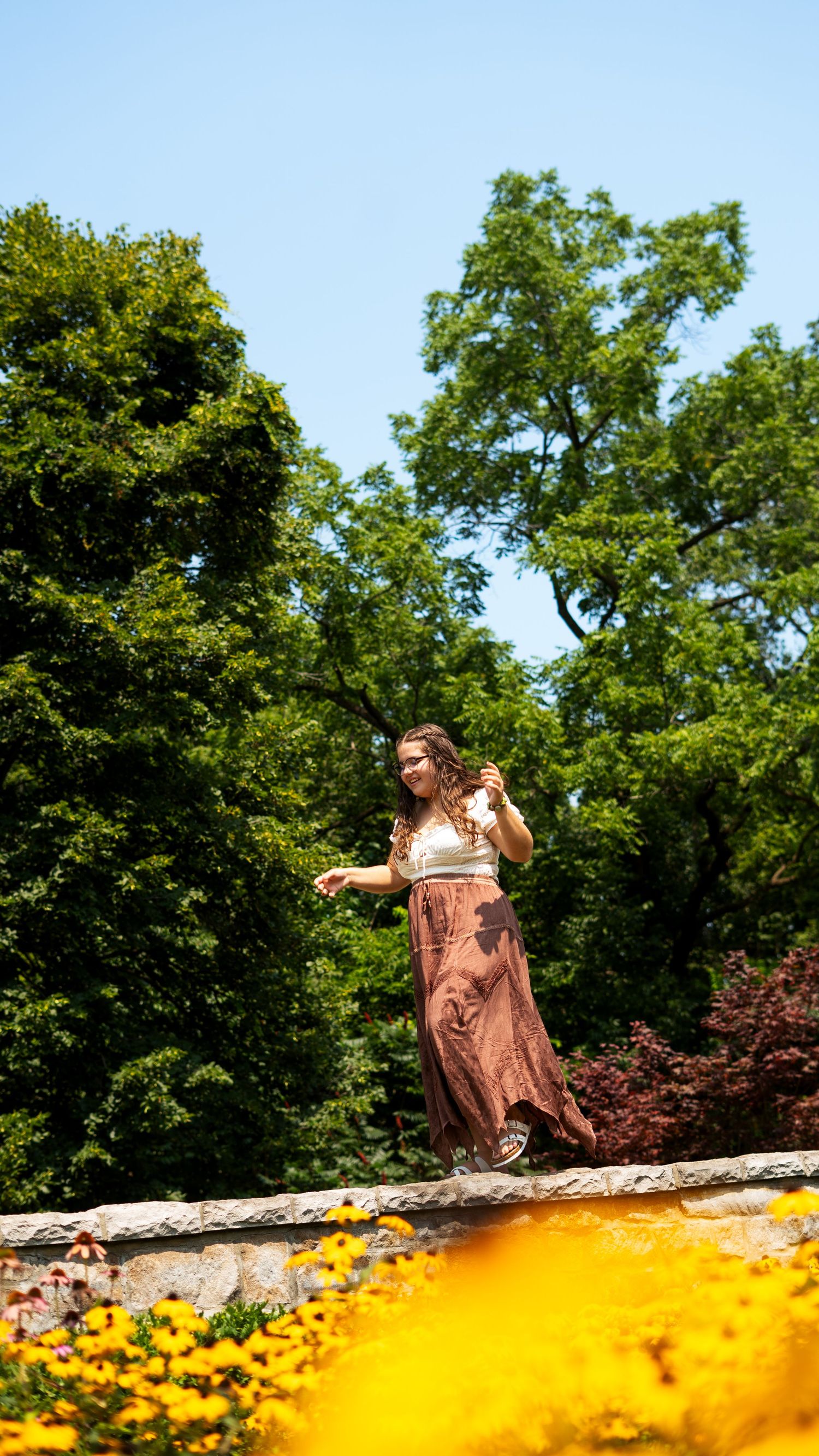 Woman in a brown dress stands on a wall against green trees with yellow flowers blooming in the foreground.