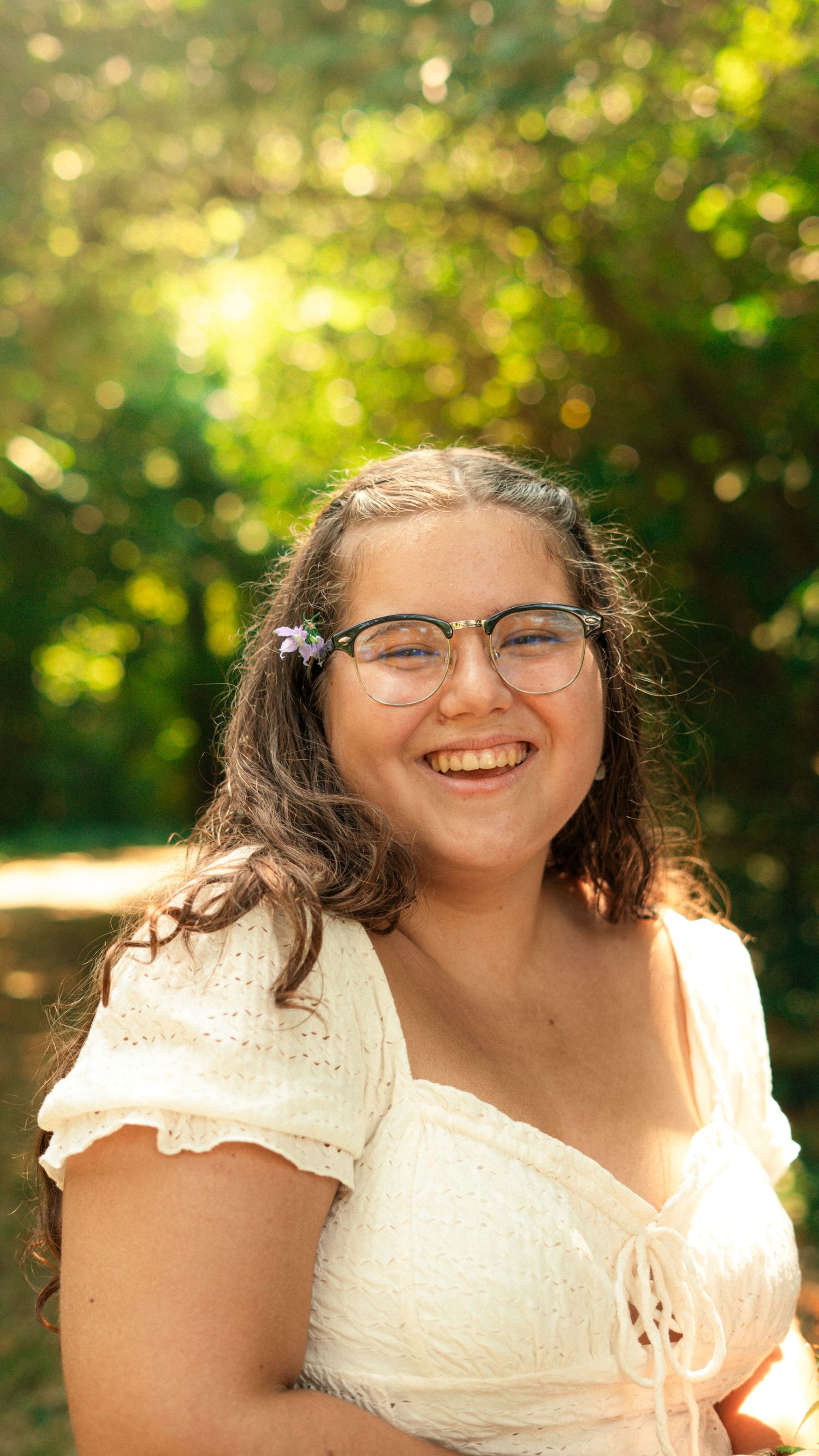 A cheerful reading session outdoors wearing a white lace top and glasses with sunlit trees in the background.