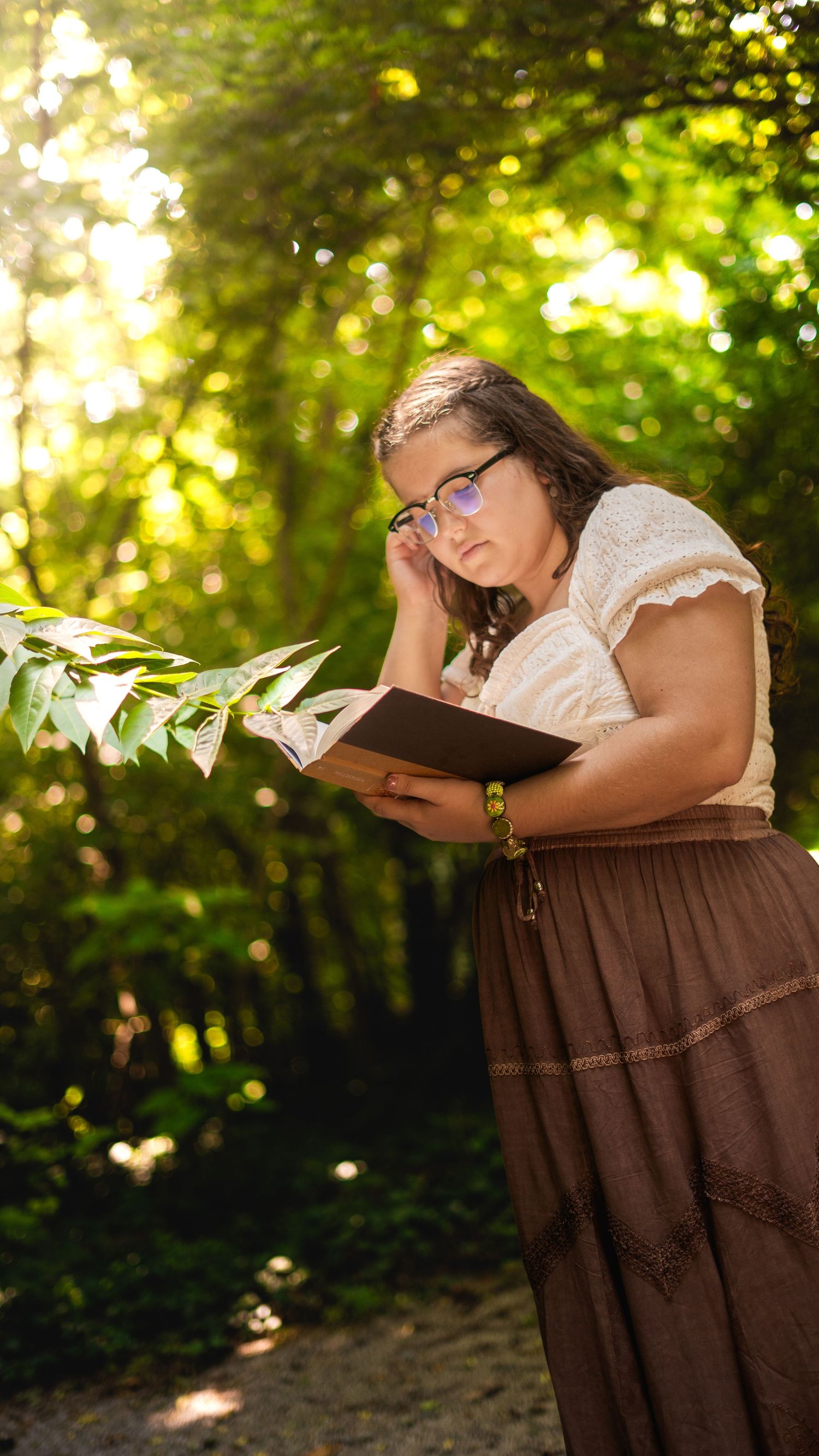 Someone reading a book while wearing a white top and brown skirt in a sunlit wooded area.