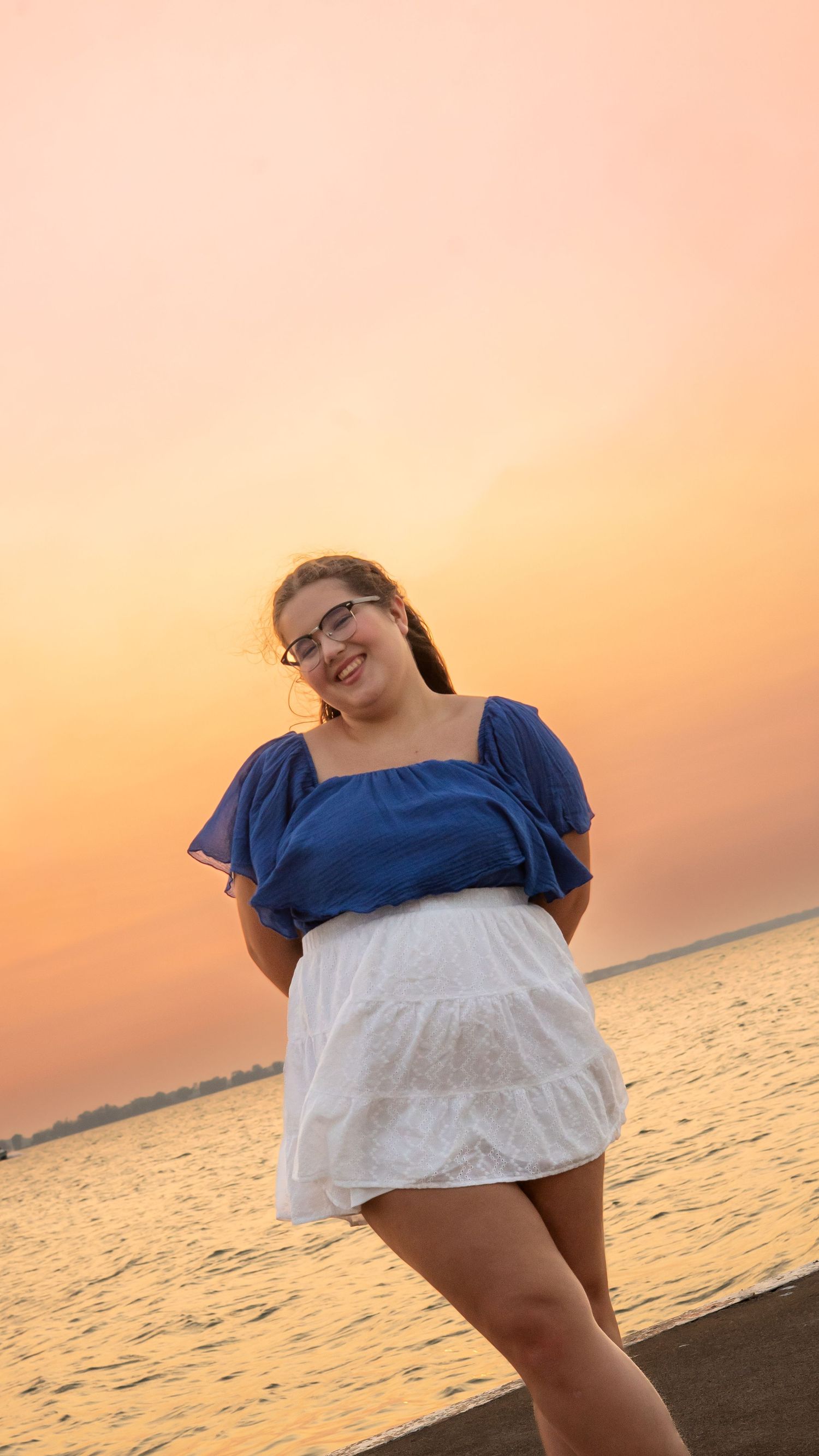 A person in a blue and white dress poses on a beach at sunset with the golden ocean waters in view.