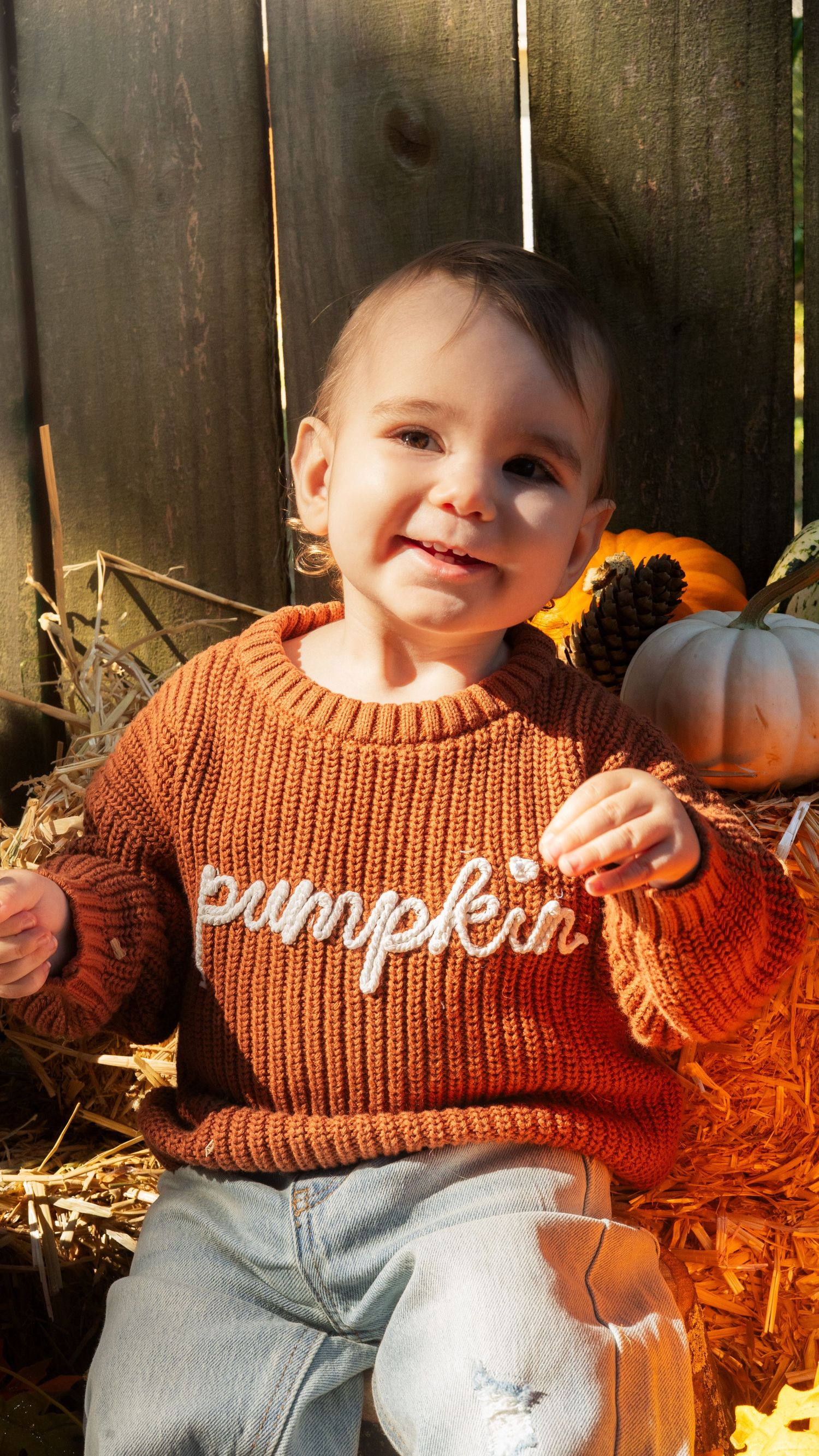Toddler in orange sweater sitting among fall decorations with small pumpkins and hay in bright autumn sunlight.
