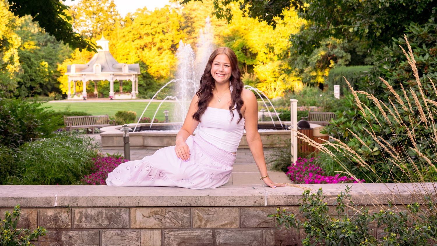 A person in a pink evening gown poses outdoors in a garden setting during golden hour lighting.