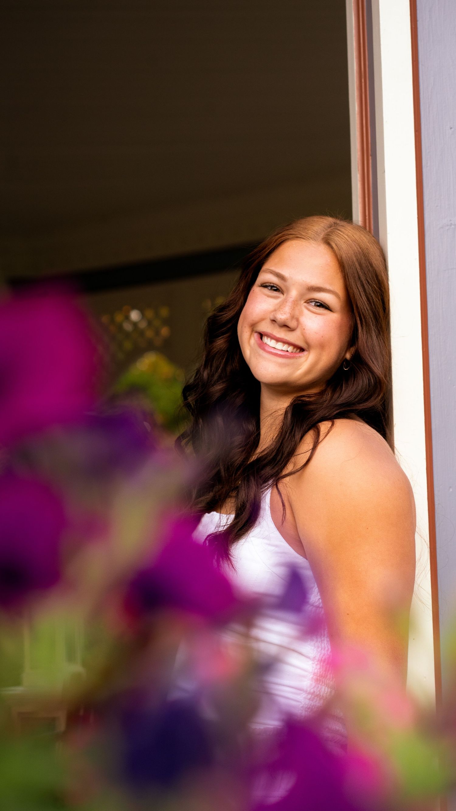 A person wearing a lavender formal gown poses against a window with blurred purple flowers in the foreground.