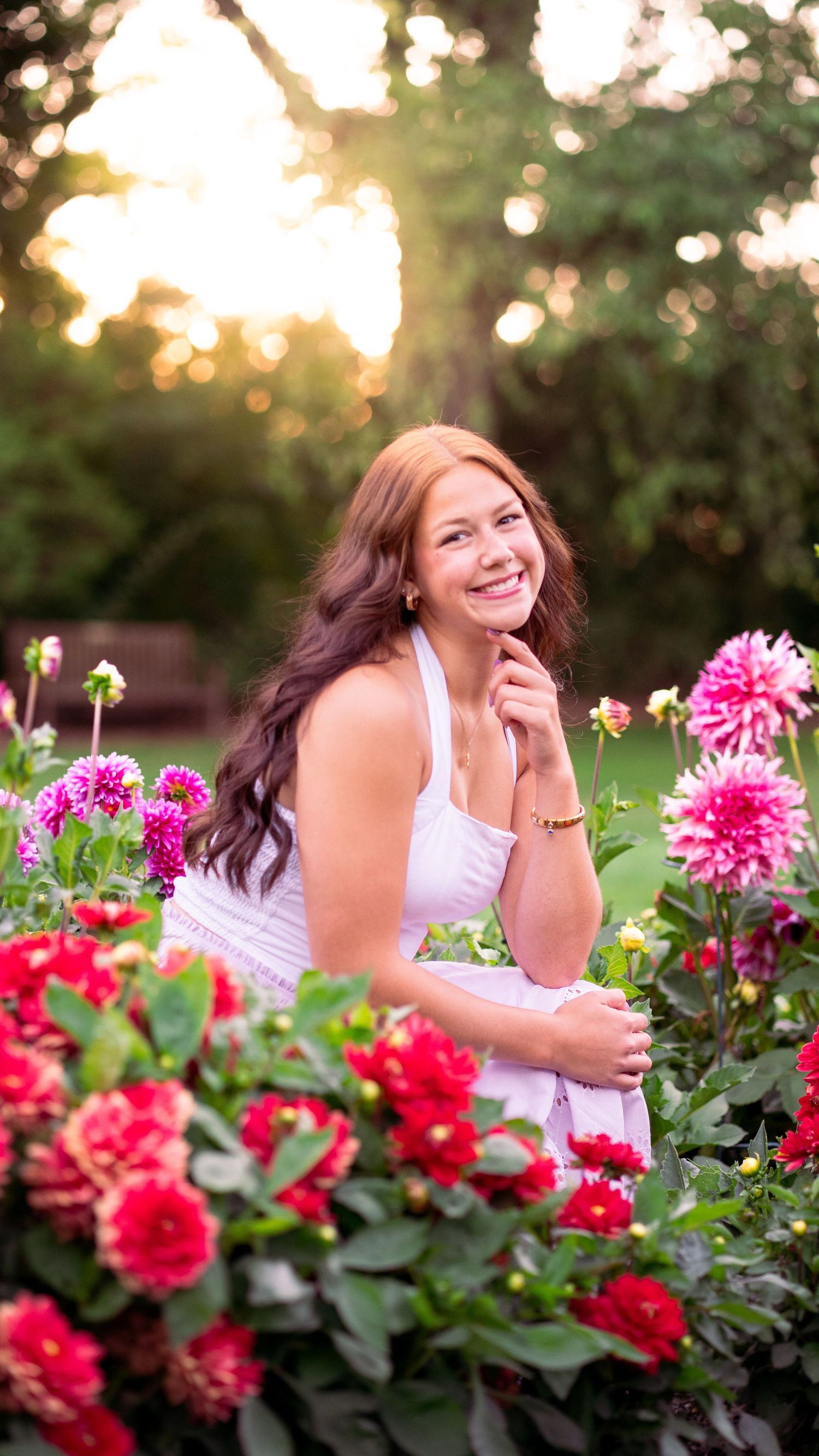 A woman in a white dress lies among vibrant pink and red dahlias during a golden sunset photoshoot.