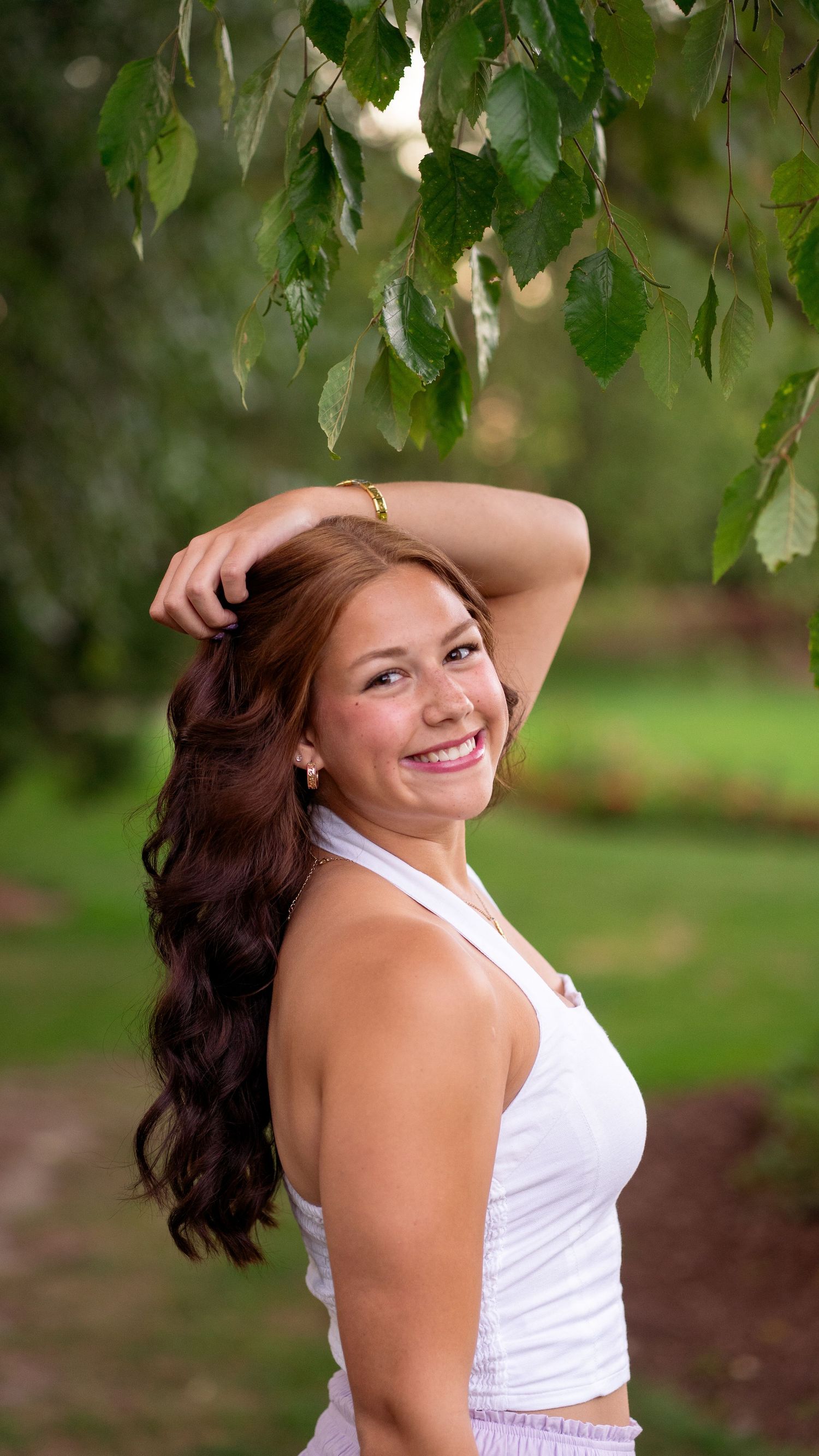 A woman in a white summer dress poses outdoors under tree branches with a bright smile.