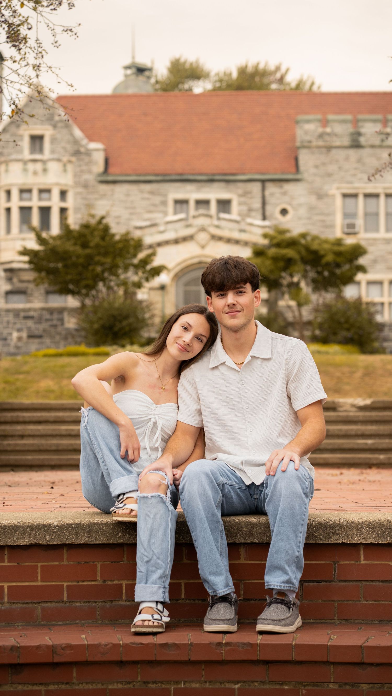 A couple sits together on brick steps in front of a grand stone building with gothic architecture on a sunny day.