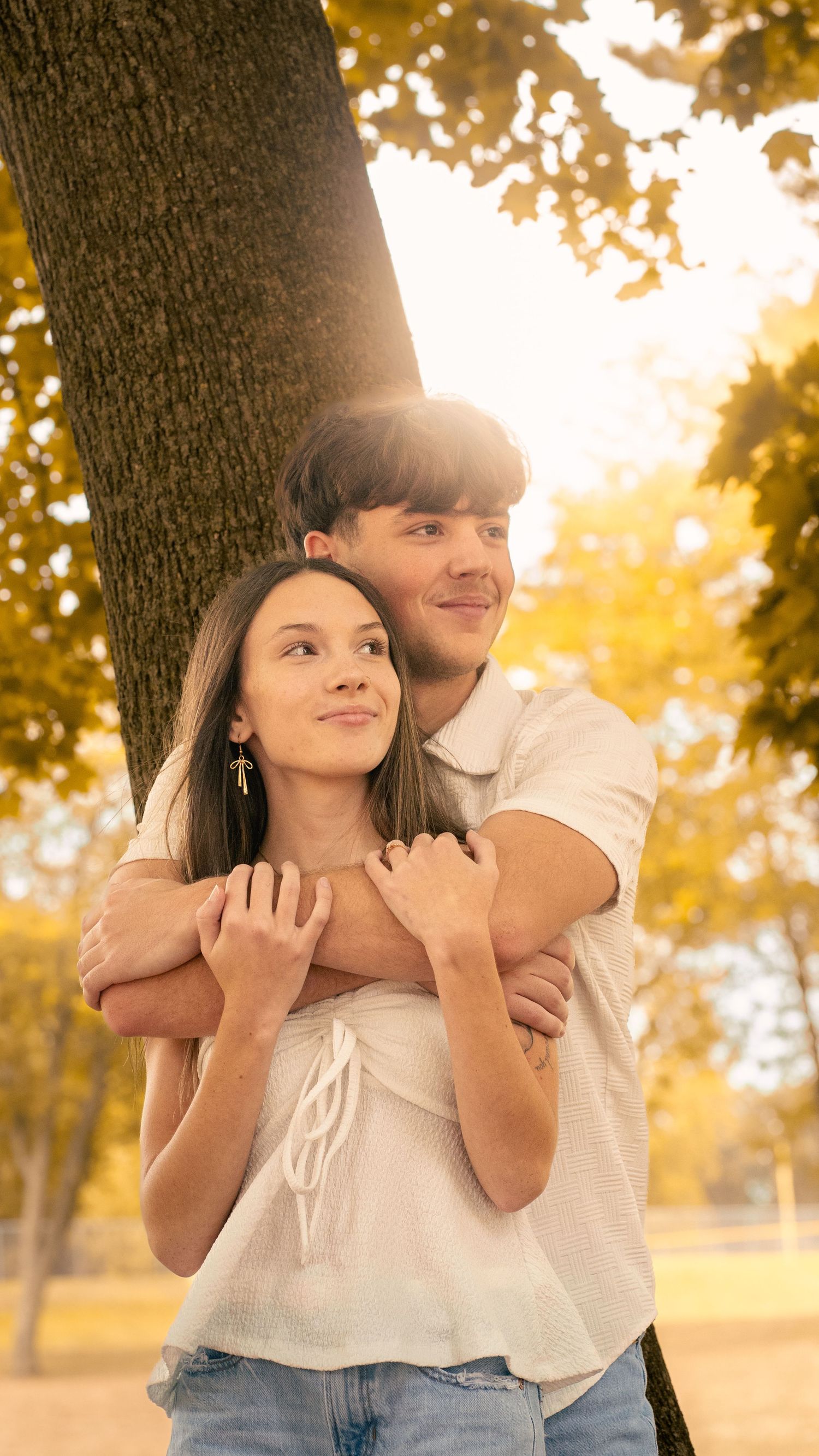 A romantic couple embraces under a tree during golden hour with autumn foliage creating a warm glow around them.