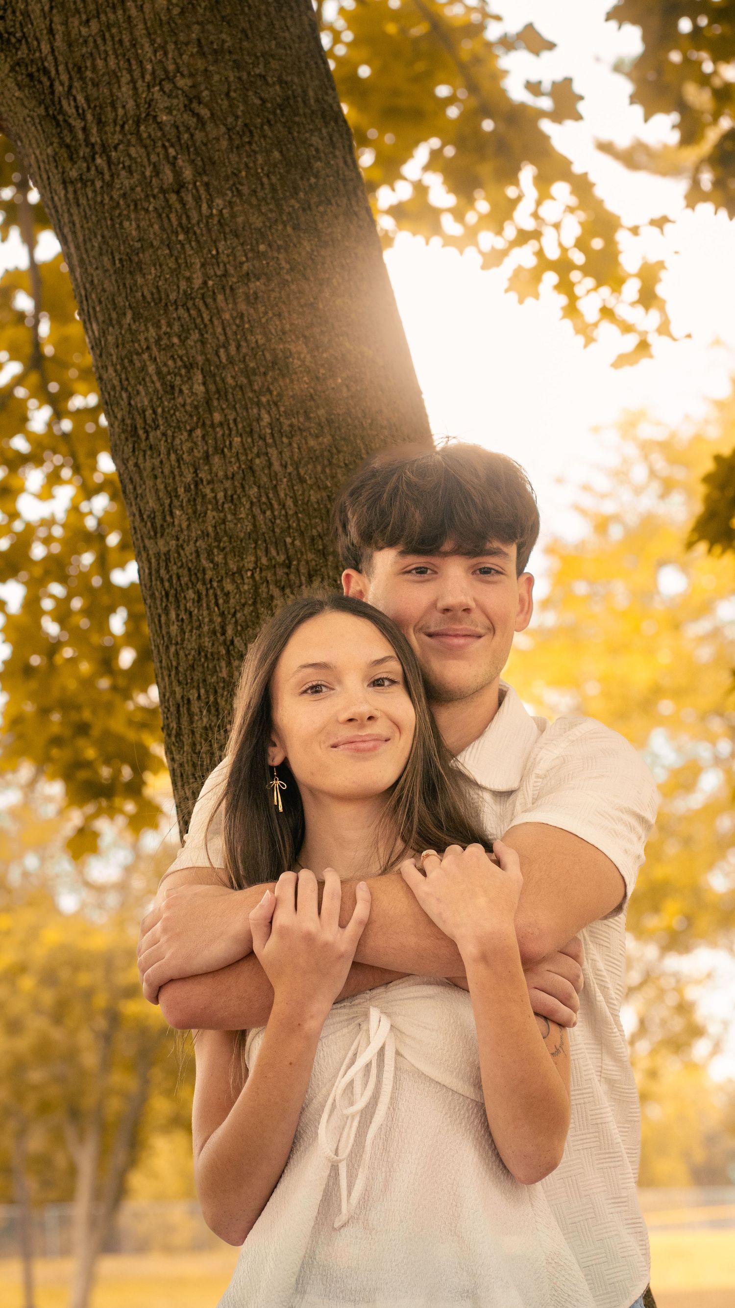 A romantic couple embraces under a tree during golden hour with autumn foliage in the background.