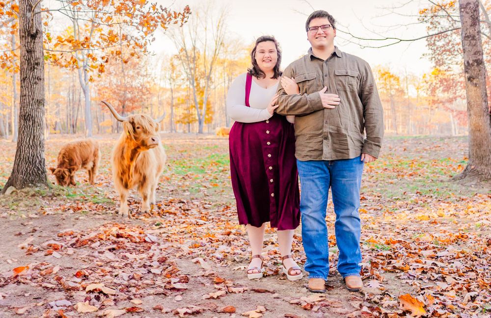 A couple stands together in fall forest with their Highland cow grazing in the background.