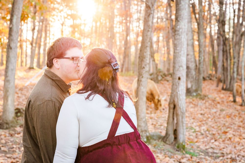 A couple embraces in a sun-dappled autumn forest with fallen leaves covering the ground.