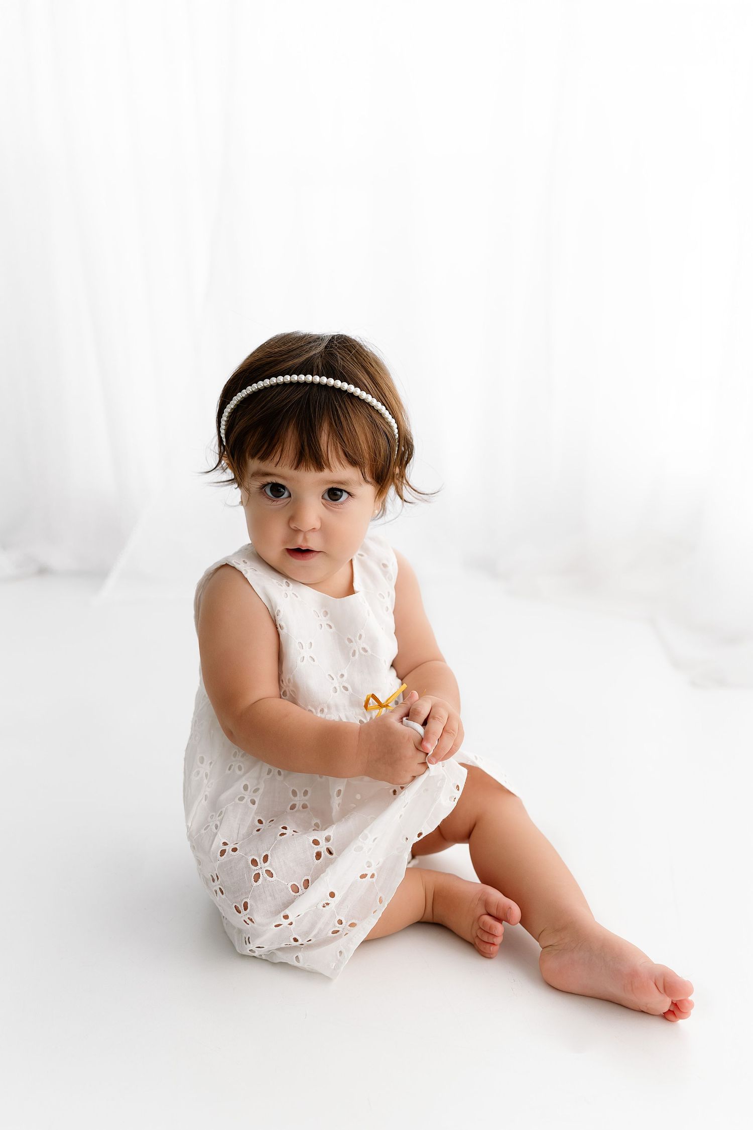 Baby in white dress sits with birthday cake and number one decoration during first birthday photoshoot.