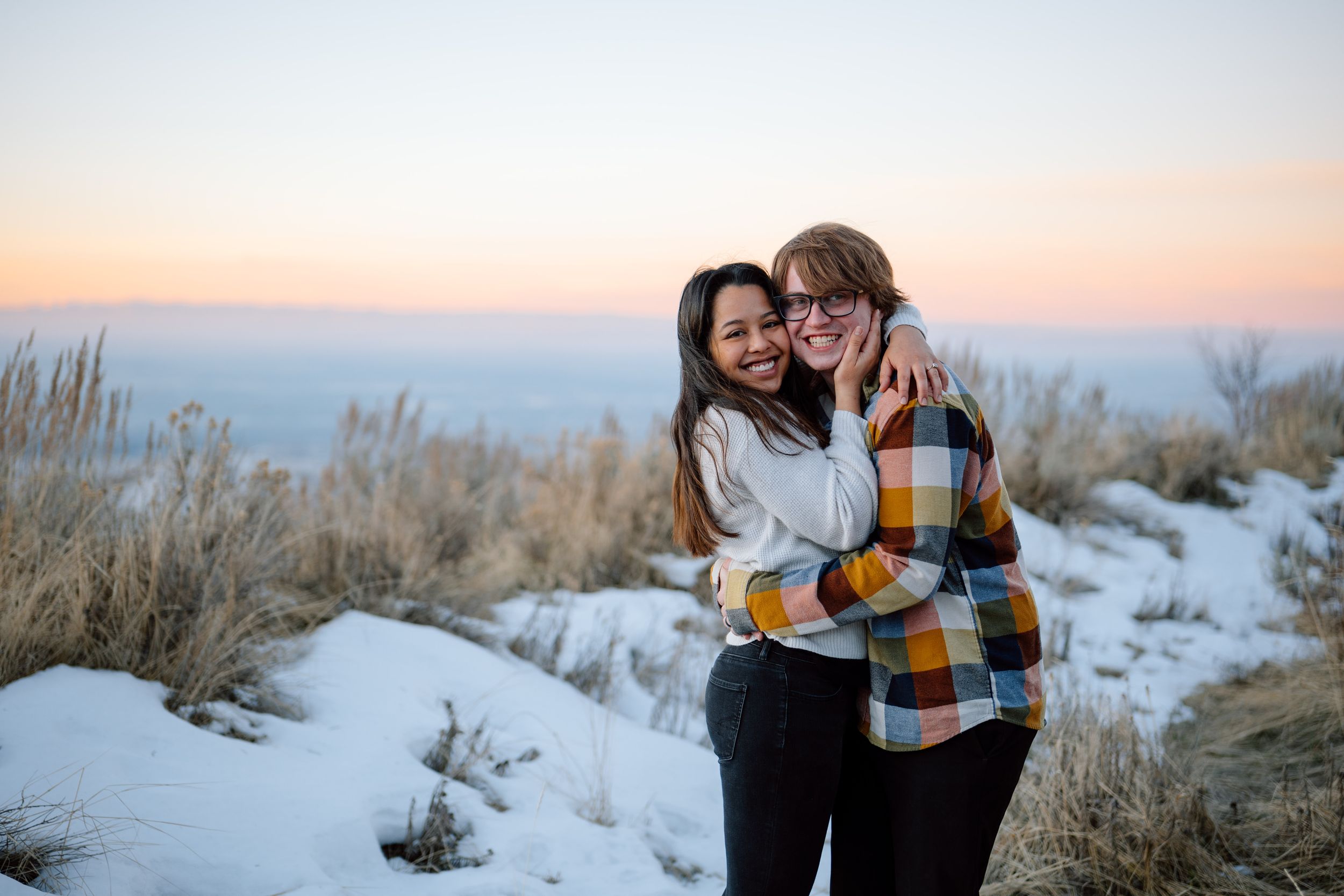 Two people embrace while standing in snowy brush during sunset against a misty mountain backdrop.