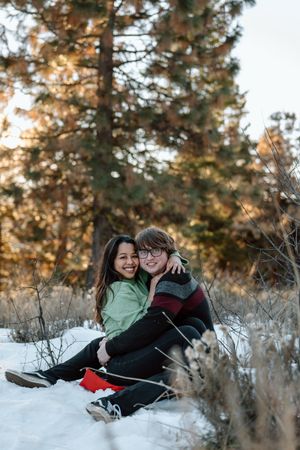 Young couple sits together on snowy ground during golden sunset with evergreen trees in background.
