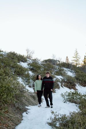 Two people walking together on a snowy mountain trail surrounded by evergreen bushes in winter.