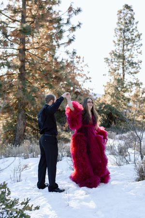 A couple in formal attire dancing in a snowy forest setting with evergreen trees, featuring a dramatic red gown.
