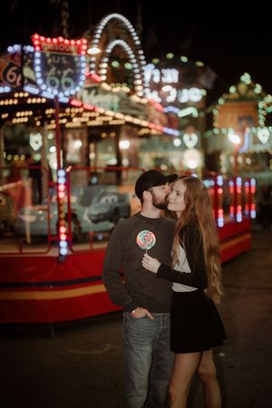 Romantic couple embraces at night in front of illuminated carnival rides at Hull Fair with colourful neon lights and decorations.
