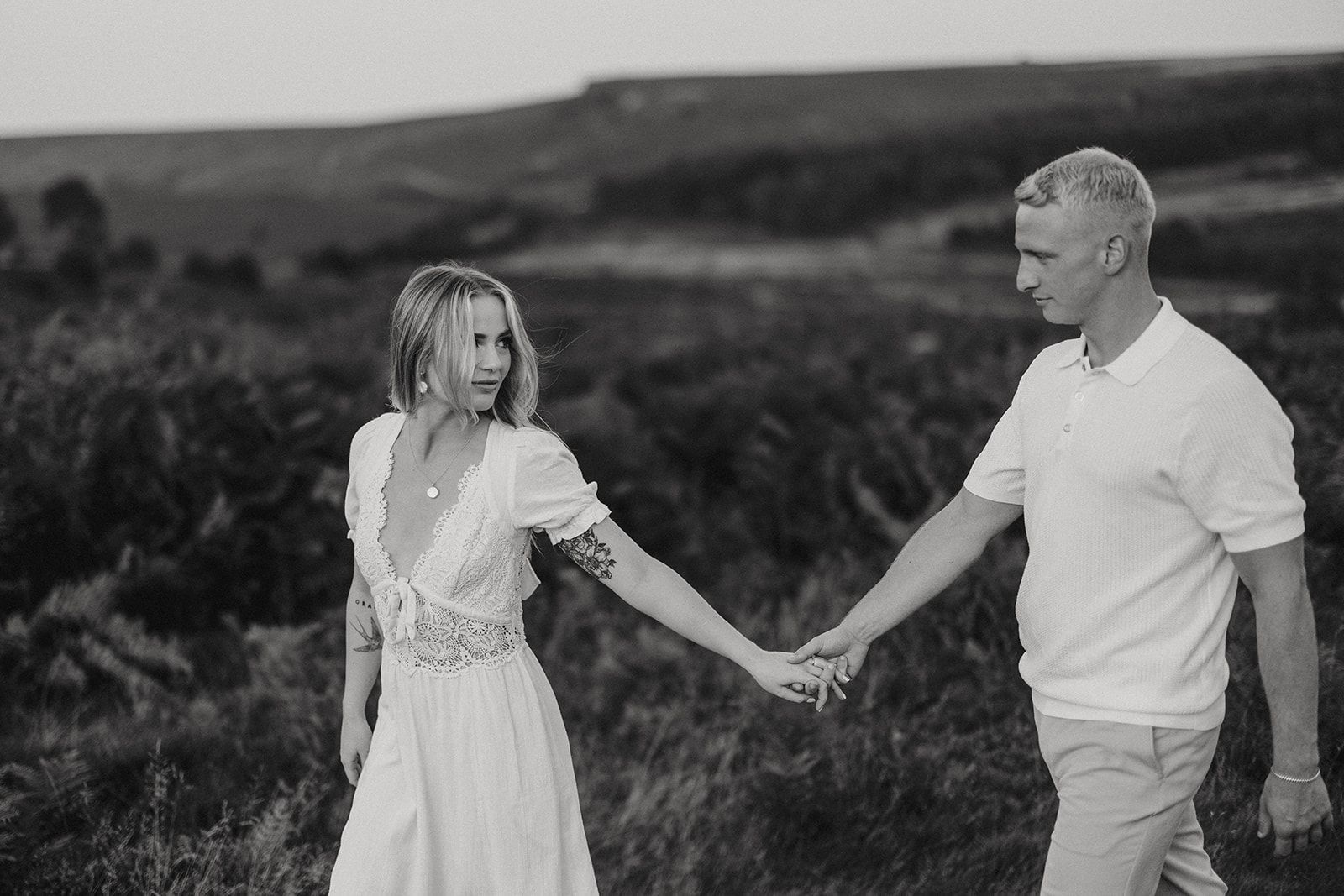 A romantic black and white photo of an engaged couple holding hands in a natural outdoor setting with rolling hills in the background.
