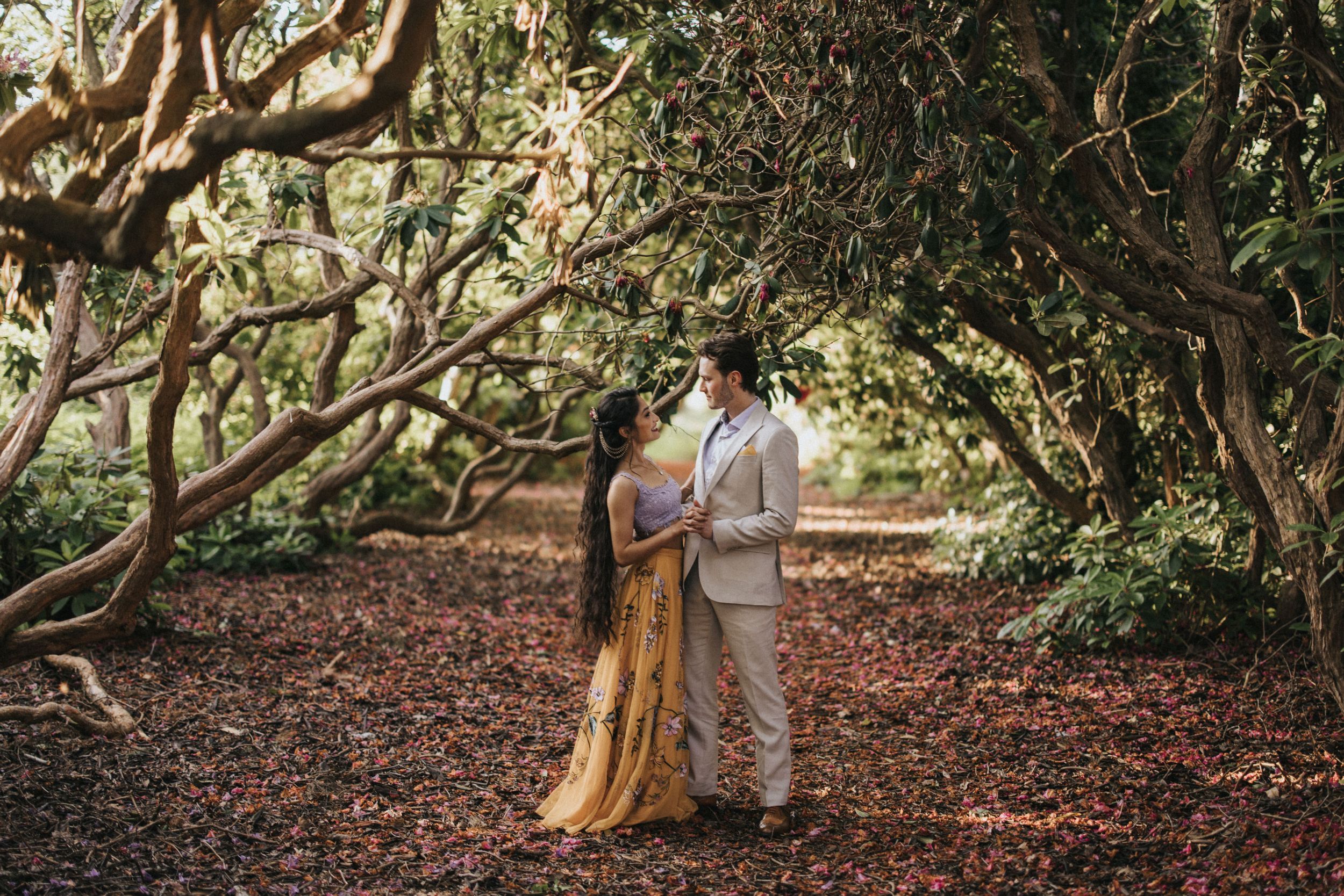 A couple stands together on a leaf-covered path beneath twisted branches of overhanging trees. Captured by Lincolnshire photographers.