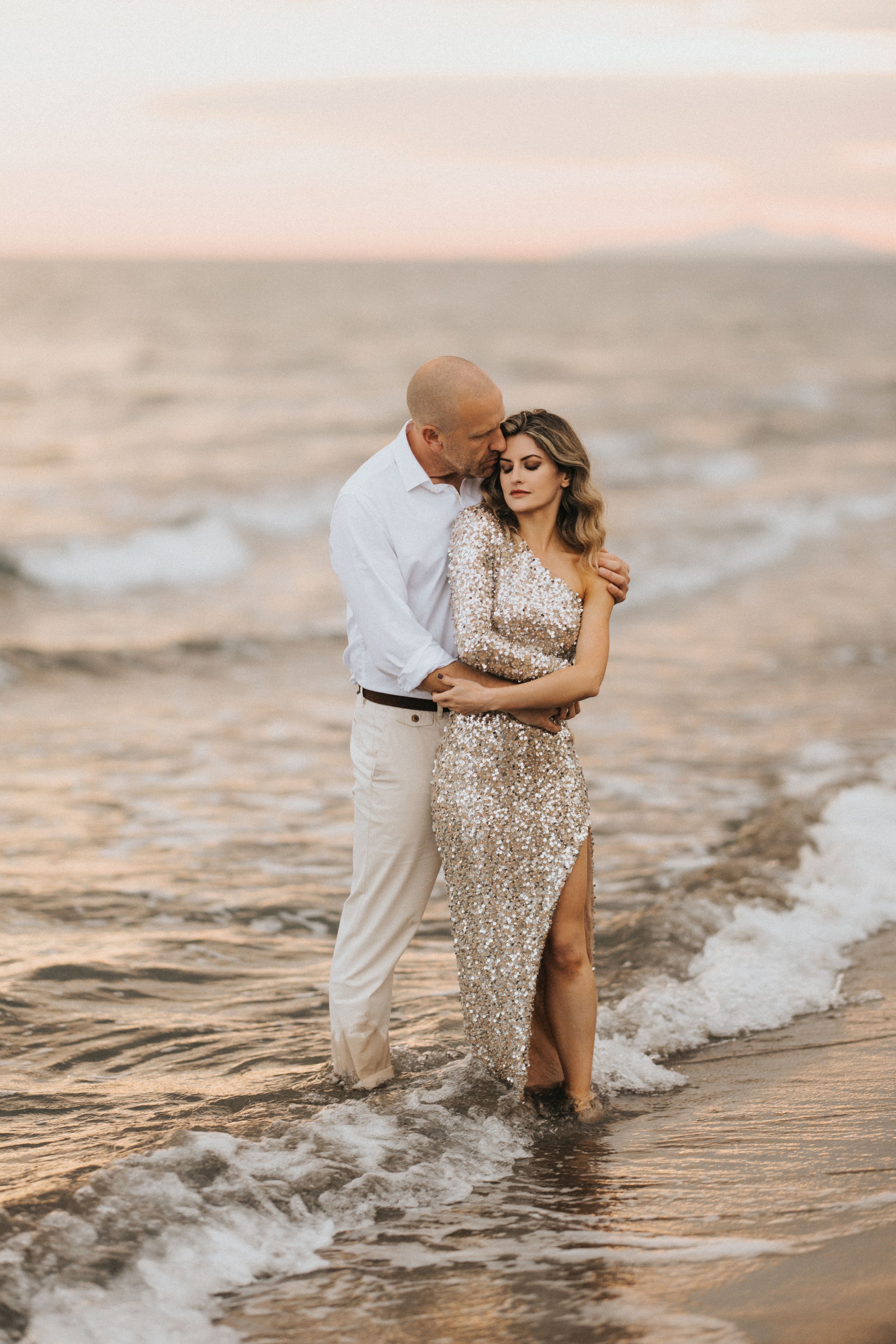 Romantic couple embracing on beach at sunset wearing formal attire as gentle waves wash over their feet.