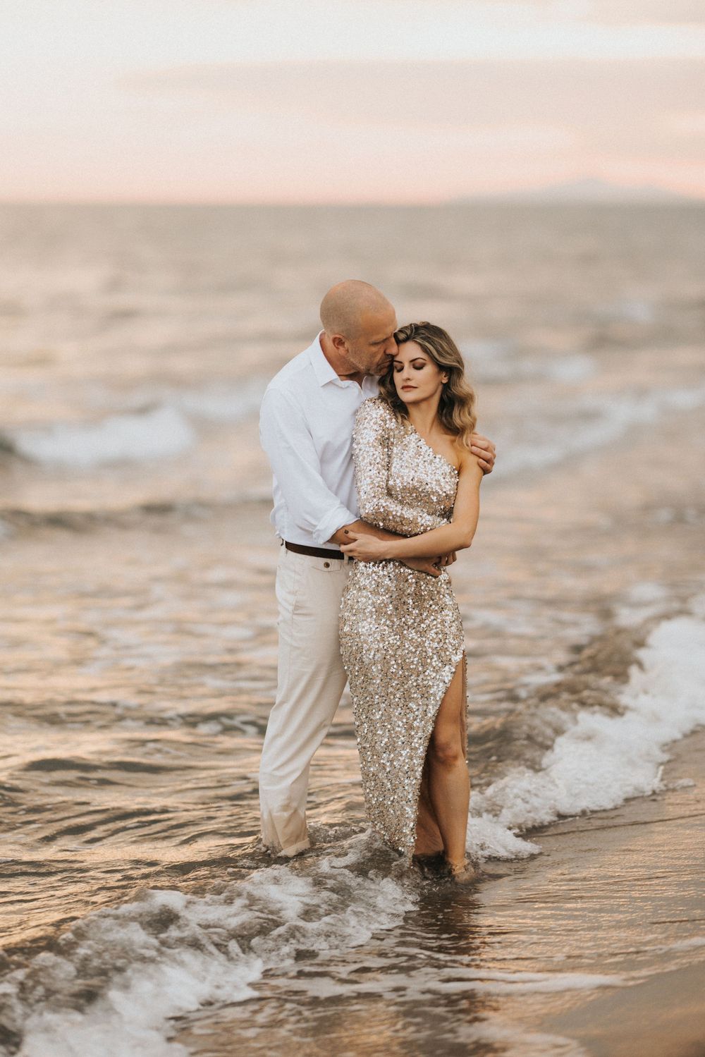 Romantic couple embracing on beach at sunset wearing formal attire as gentle waves wash over their feet.
