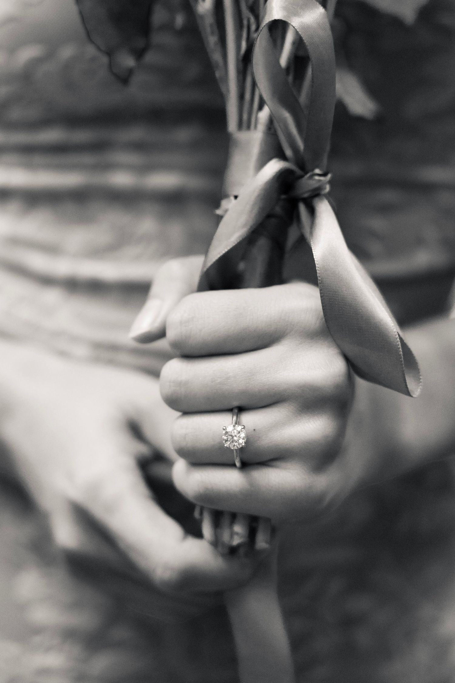 Black and white close-up of hands holding a ribbon bow, showcasing a diamond engagement ring.