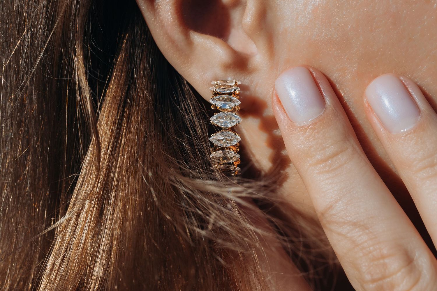 Close-up of sparkling diamond stud earring being worn with neutral manicured nails and brown hair visible in frame.