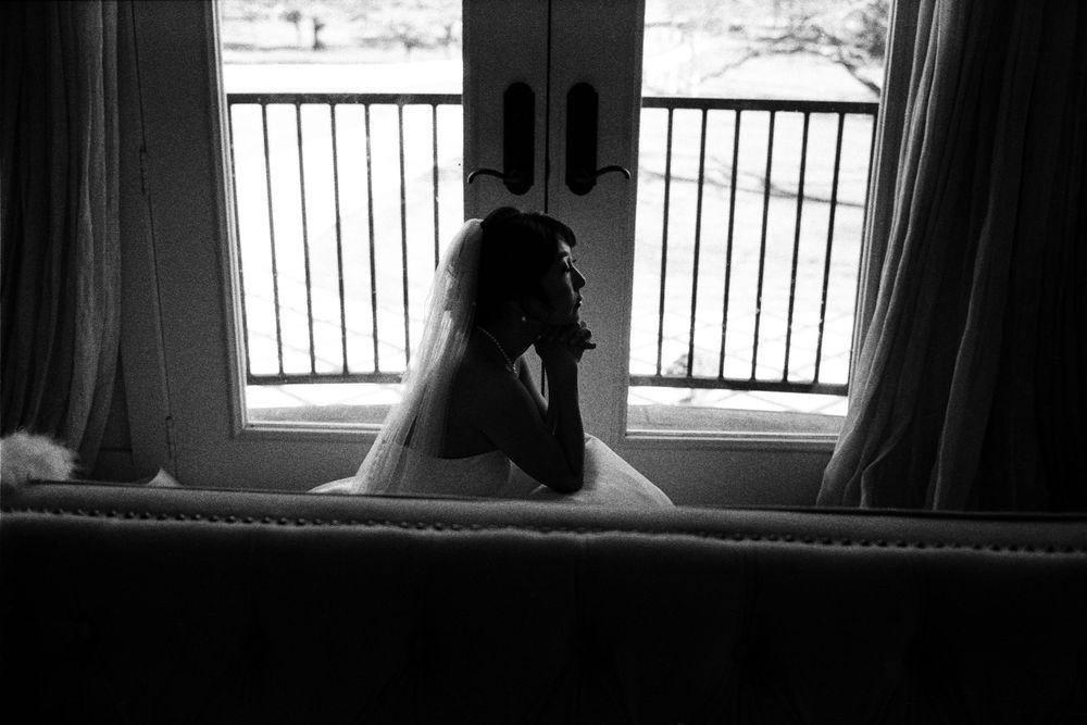 Silhouette of a bride in a wedding gown sitting pensively by a window with French doors in black and white.