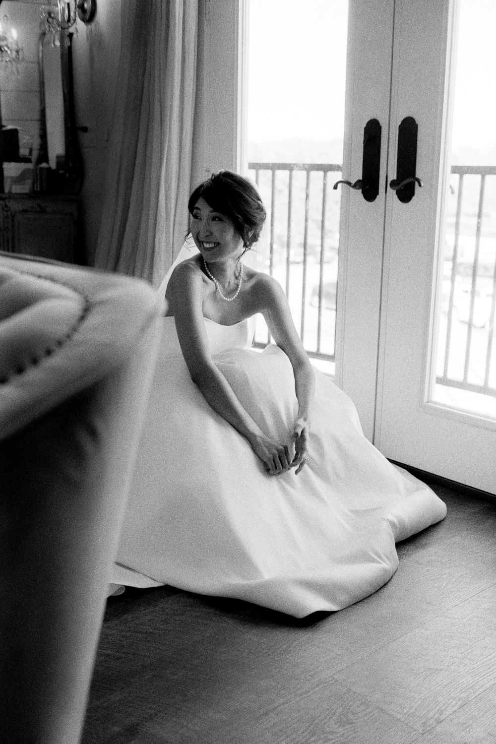 Elegant bride in a strapless white gown sitting on the floor near French doors, smiling softly in black and white.