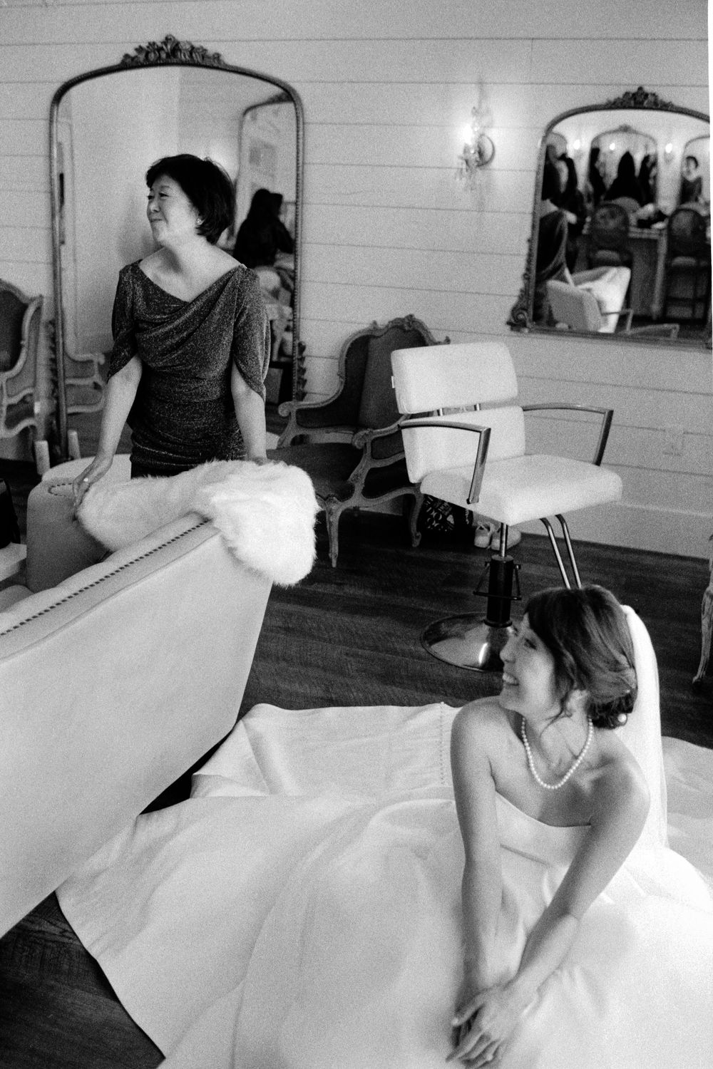 Bride in white gown and woman in gray dress share a candid moment in a vintage salon with ornate mirrors.